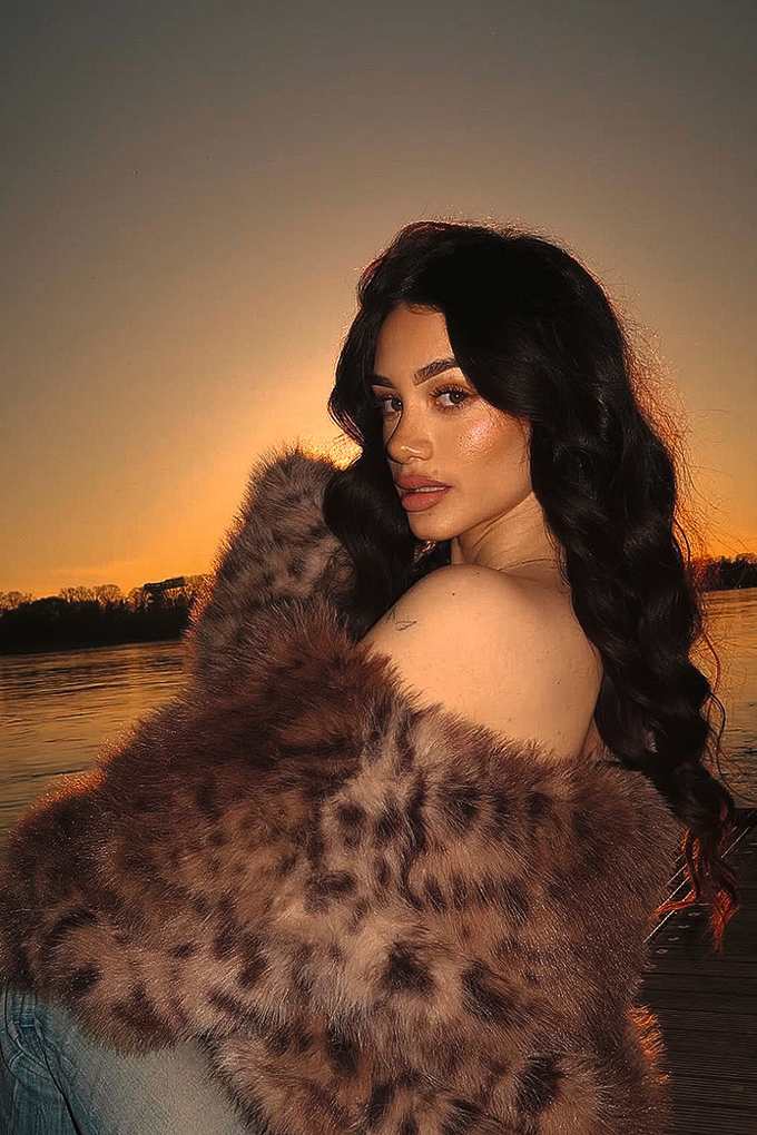 Influencerin Rossella posiert im schulterfreien Fake Fur-Mantel vor Sonnenuntergangs-Kulisse