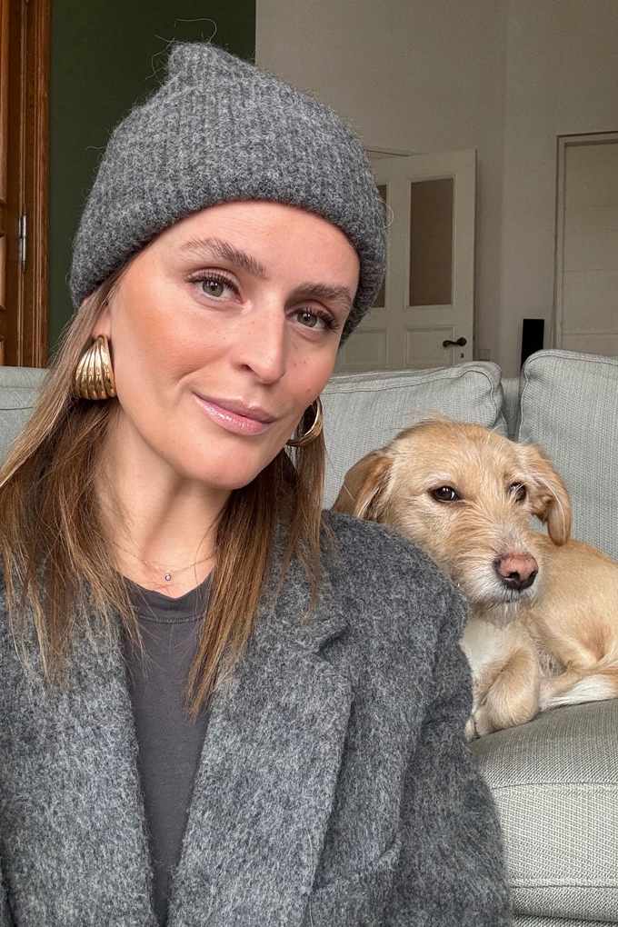 Frau in grauem Mantel mit passender Mütze sitzt vor Couch, Hund liegt daneben