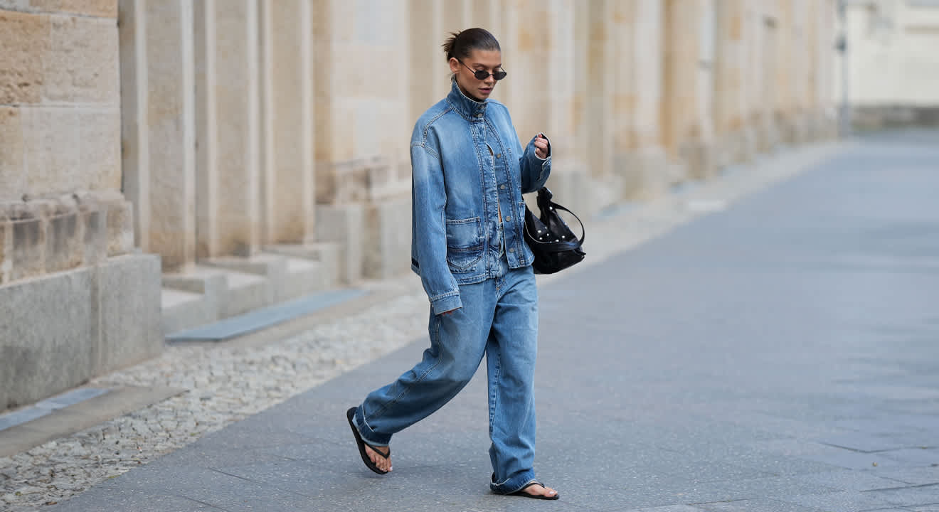 Frau in komplettem Jeans-Outfit, Jacke und Hose im Denim-Look, mit offenen Sandalen, in urbaner Kulisse