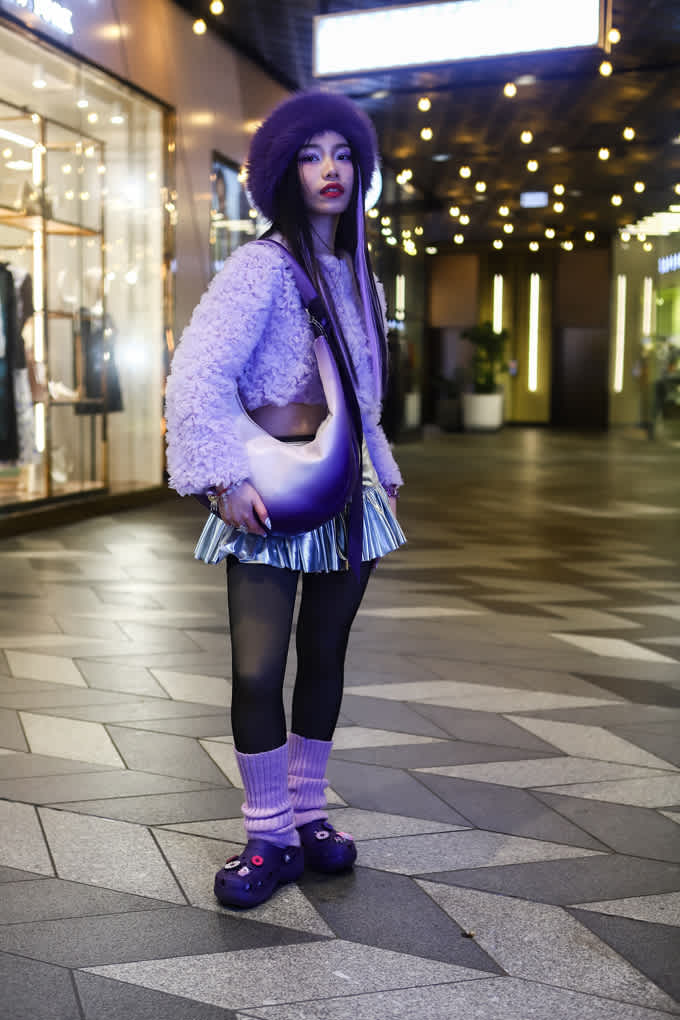 Frau in lila Plüschjacke und passender Halfmoon Bag, trägt Metallic-Minirock in Einkaufsgasse