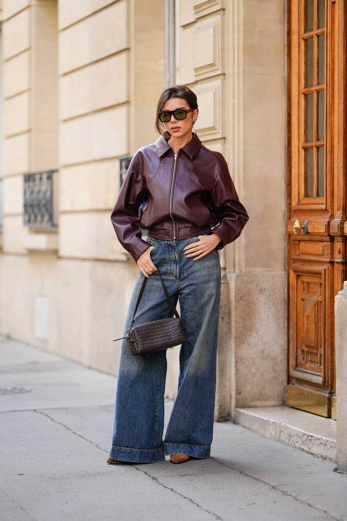 Frau in dunklem Bordeaux-Blouson, kombiniert mit Wide Leg-Pants und geflochtener Schultertasche
