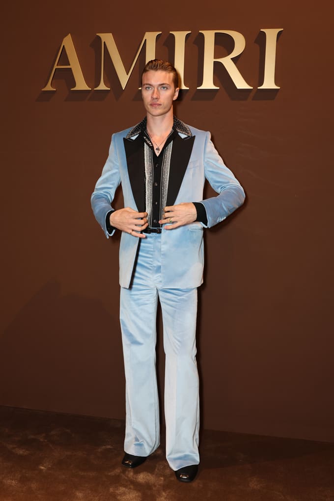 Mann trägt hellblaue Anzugkombi mit Hahnentrittjacke vor "Amiri"-Schriftzug an Wand