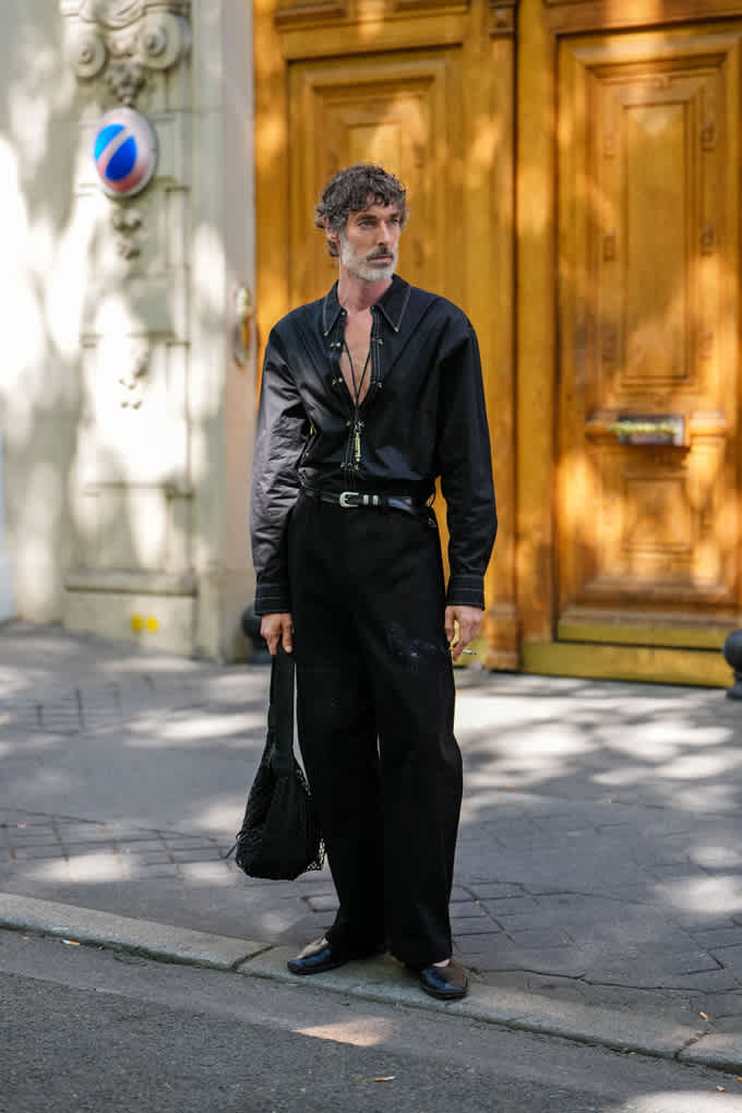 Mann steht an Straßenrand, trägt schwarze Hose und passendes Hemd mit tief geöffnetem Kragen