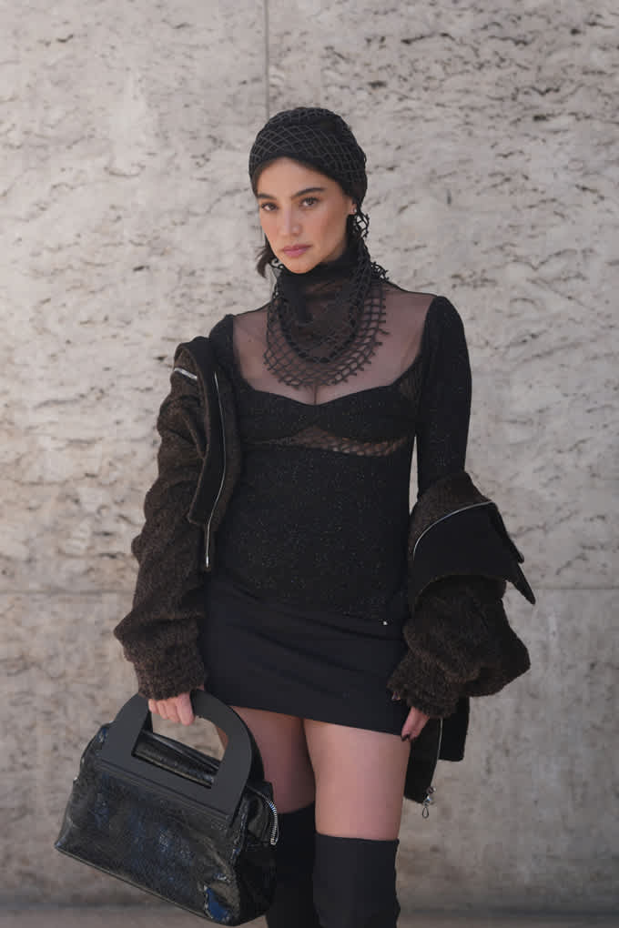 Frau in schwarzem Grunge-Outfit, trägt schwarze Handtasche, stehend vor Betonwand