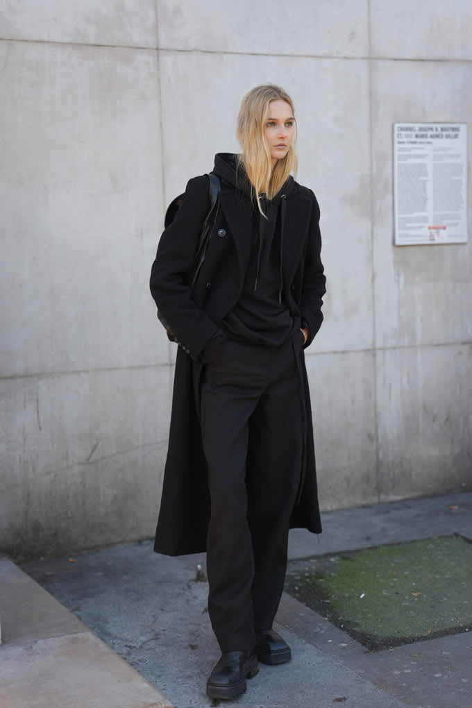 Blonde Frau trägt langen schwarzen Mantel im Grunge-Style, kombiniert mit schwarzer Hose und Stiefeln