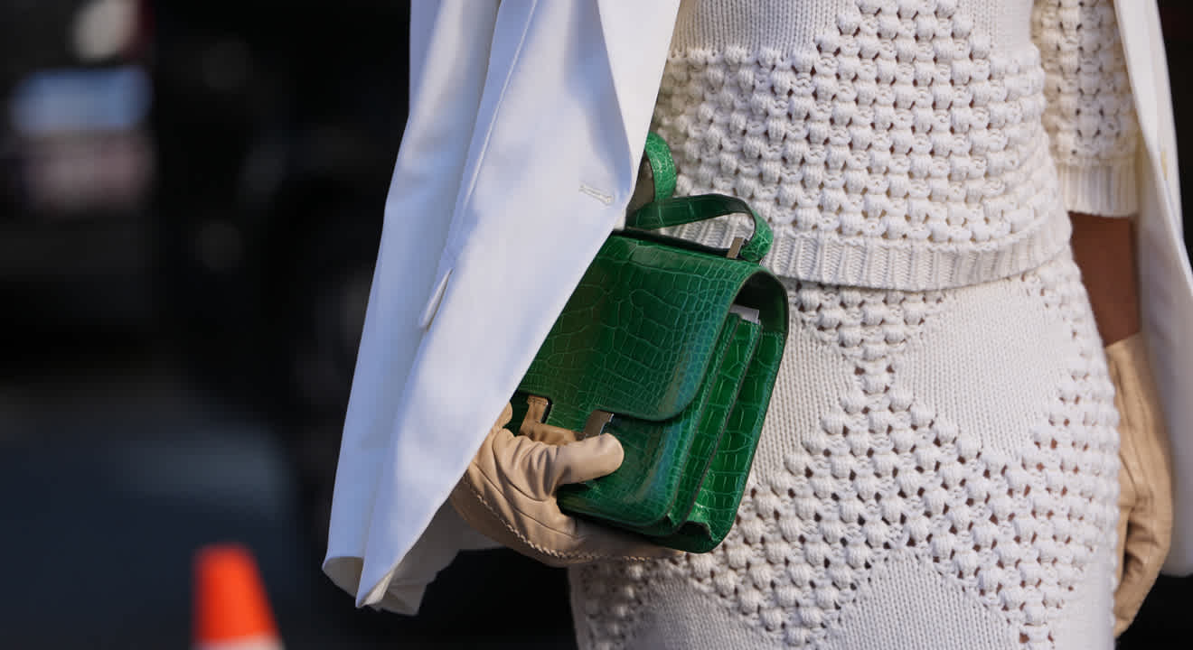 Frau in weiß-strukturiertem Strickoutfit, mit weißem Blazer, hält grüne Handtasche in der Hand