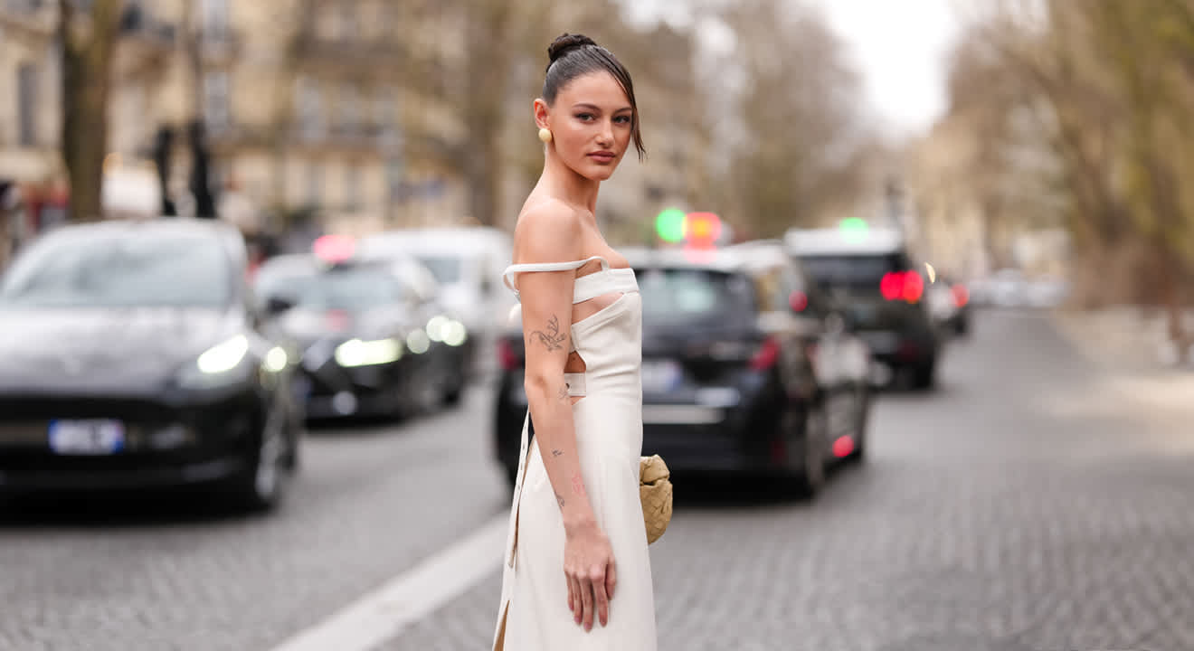 Frau mit Dutt, trägt weißes schulterfreies Kleid, steht inmitten einer Straße umgeben von Autos