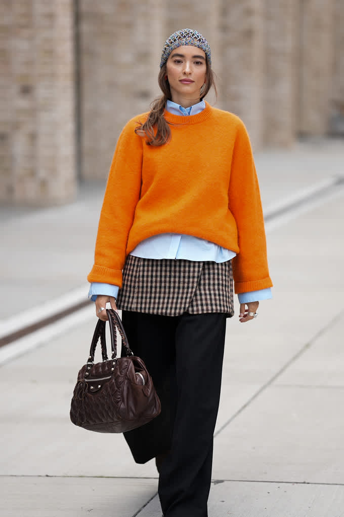 Frau trägt Layering-Look aus orangefarbenem Pullover mit weißem Shirt, Karo-Top und schwarzer Hose
