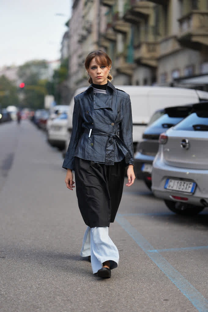 Frau auf Straße trägt asymmetrischen Trenchcoat in Grau, kombiniert mit weiter Hose und Rock im Layering-Look