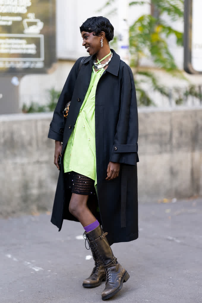 Frau mit schwarzen Haaren trägt grünes Kleid unter schwarzem Oversize-Trenchcoat, kombiniert mit Boots
