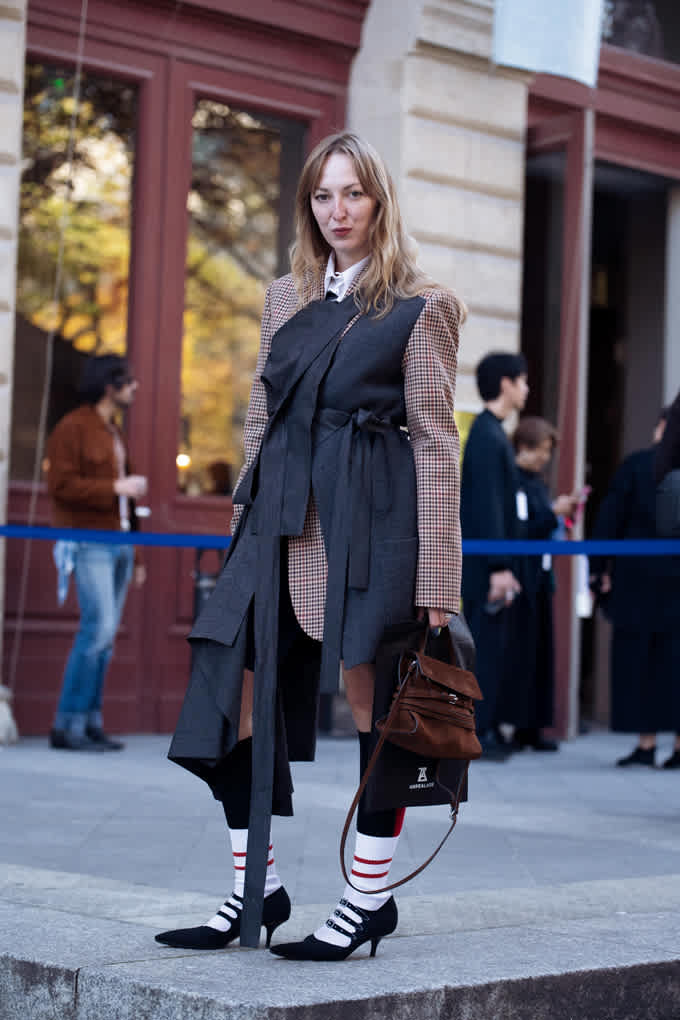 Blonde Frau steht auf Gehweg, trägt Layering-Outfit im Dark Academia-Style