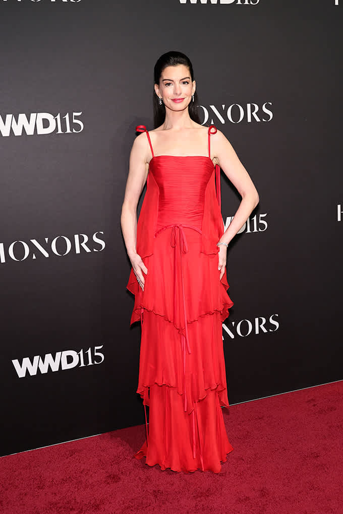 Anne Hathaway steht in knallrotem Chiffonkleid auf rotem Teppich, vor Logo-Wand