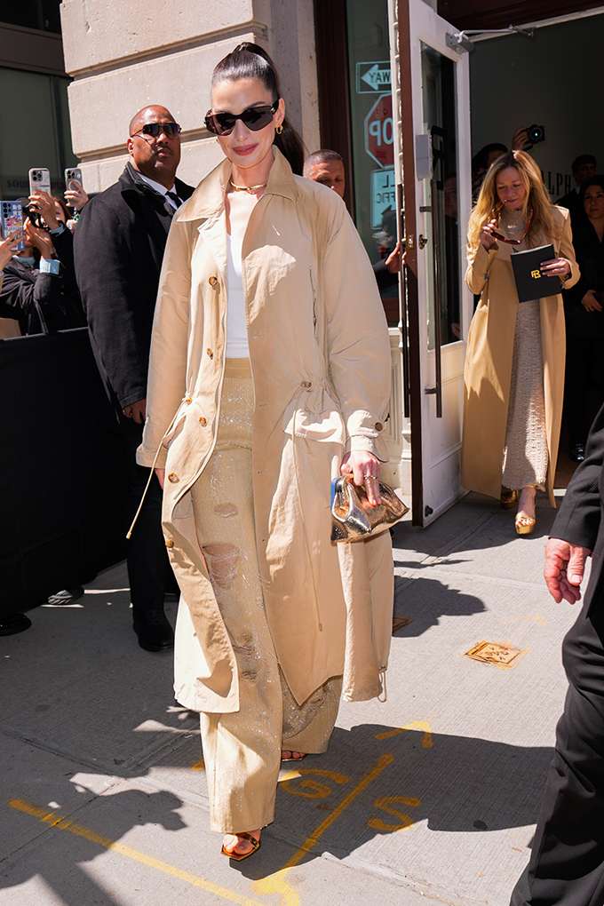 Anne Hathaway in beigem Trenchcoat bei Event, trägt goldene Mini-Clutch und schwarze Sonnenbrille