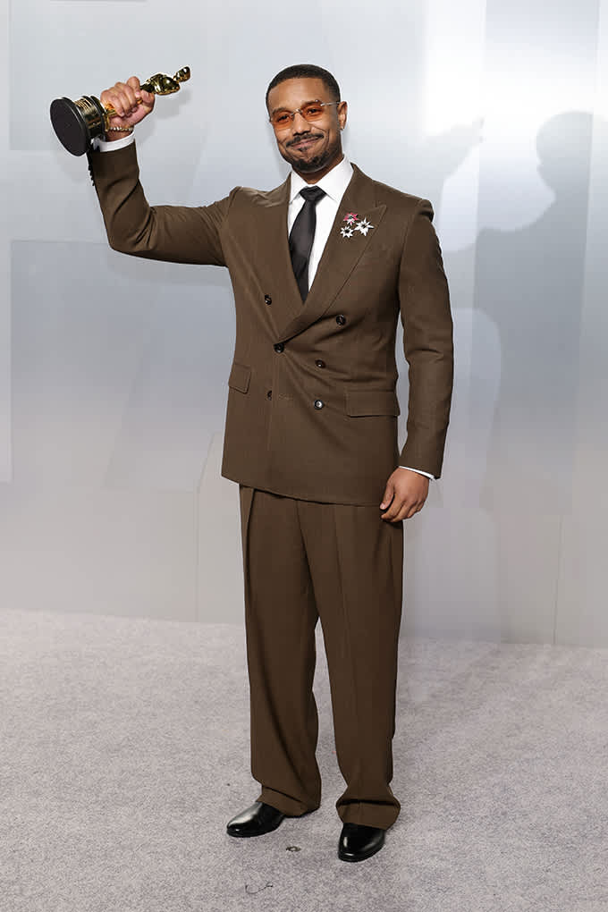 Michael B. Jordan in schlicht-braunem Zweireiher-Anzug, hält Oscar in der rechten Hand