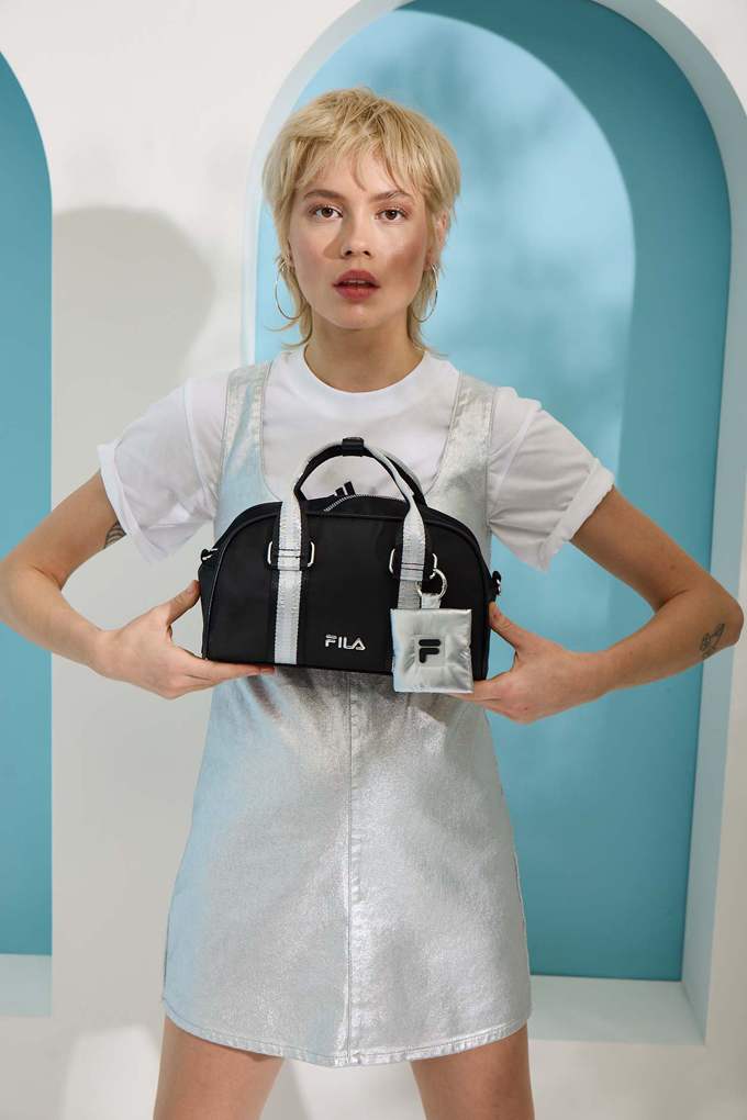 Blonde Frau in weißem Basic-Kleid hält schwarz-weiße FILA-Handtasche vor der Brust