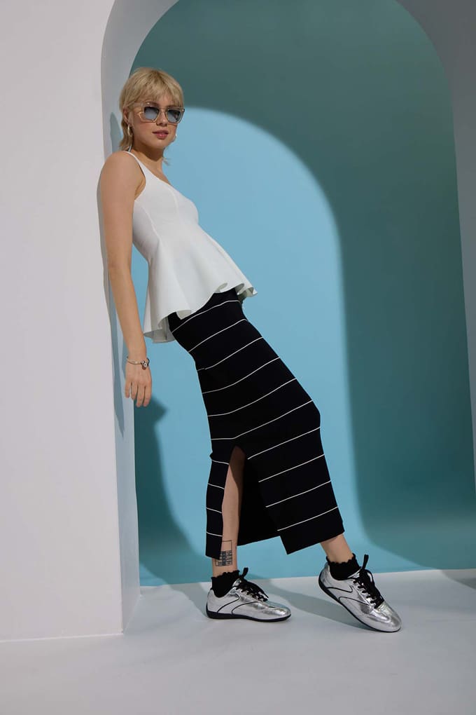 Frau in weißem Peplum-Top, trägt Midi-Rock und silberne Sneaker, lehnt an Steinbogen