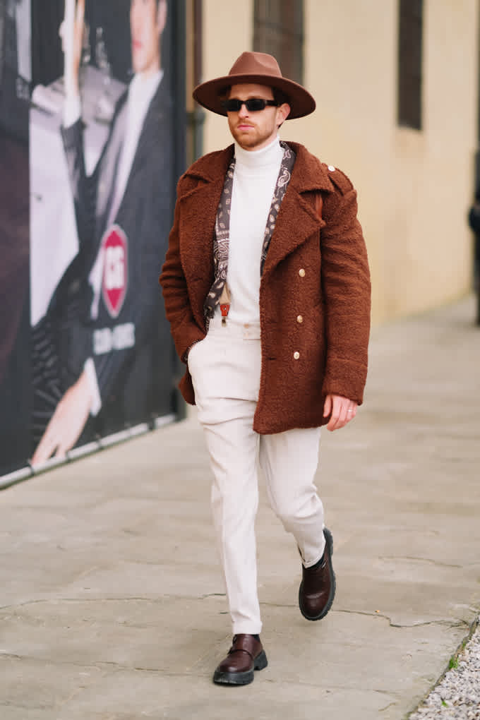Mann in braunem Teddy-Coat, kombiniert mit weißer Hose und Hemd, mit braunem Hut