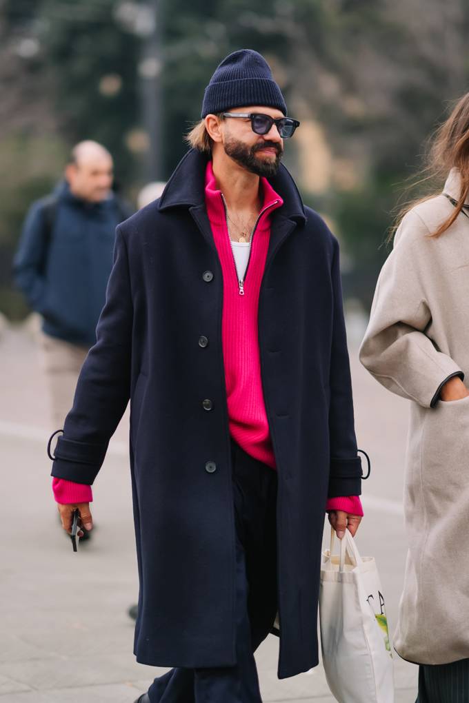 Männliches Model trägt schwarzen Mantel in Kombination mit knallpinkem Zip-Pullover und Beanie