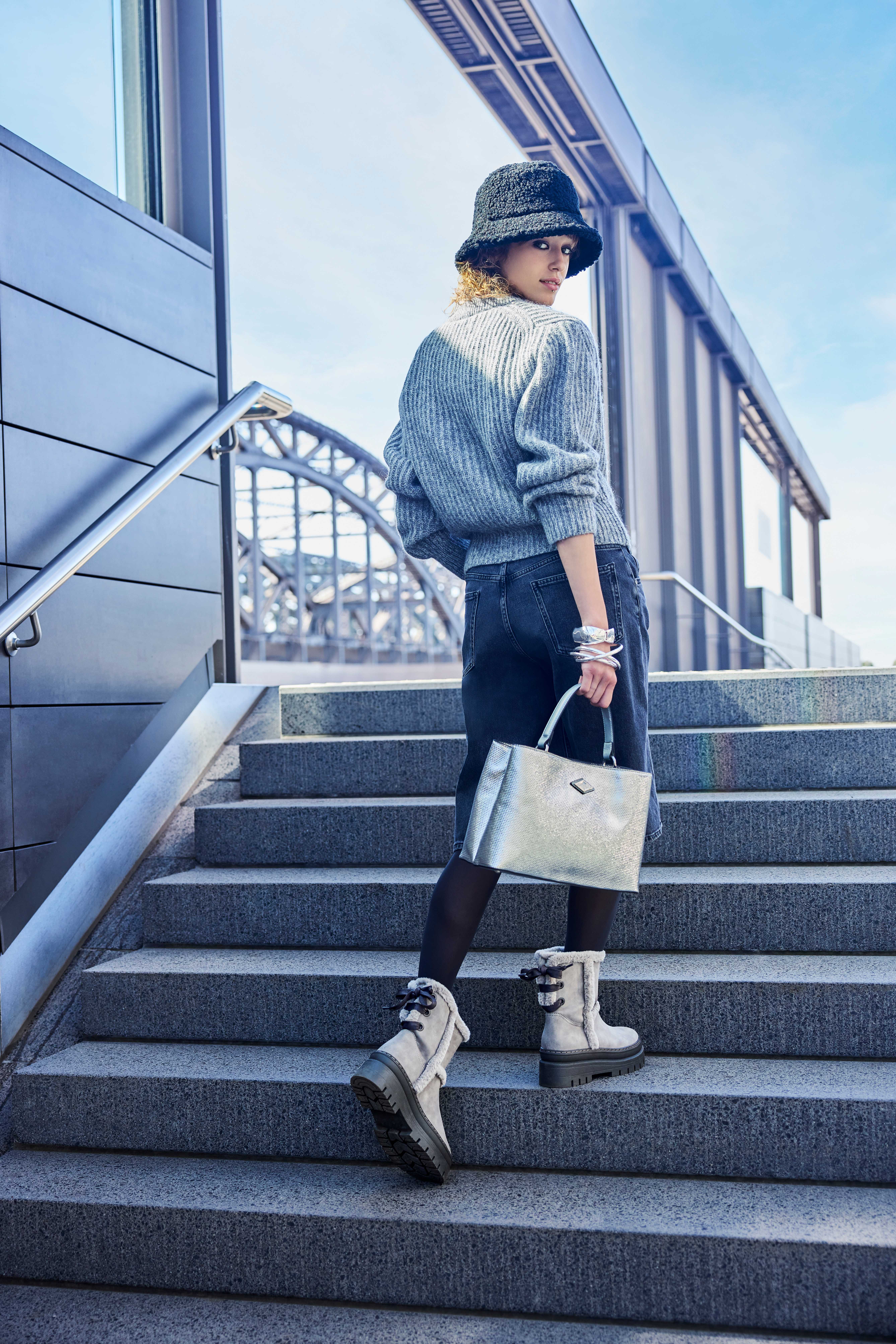 Frau in grauem Pullover, mit schwarzem Jeansrock, beigen Winterstiefeln und schwarzem Bucket Hat, gehend auf einer Treppe mit silberner Handtasche