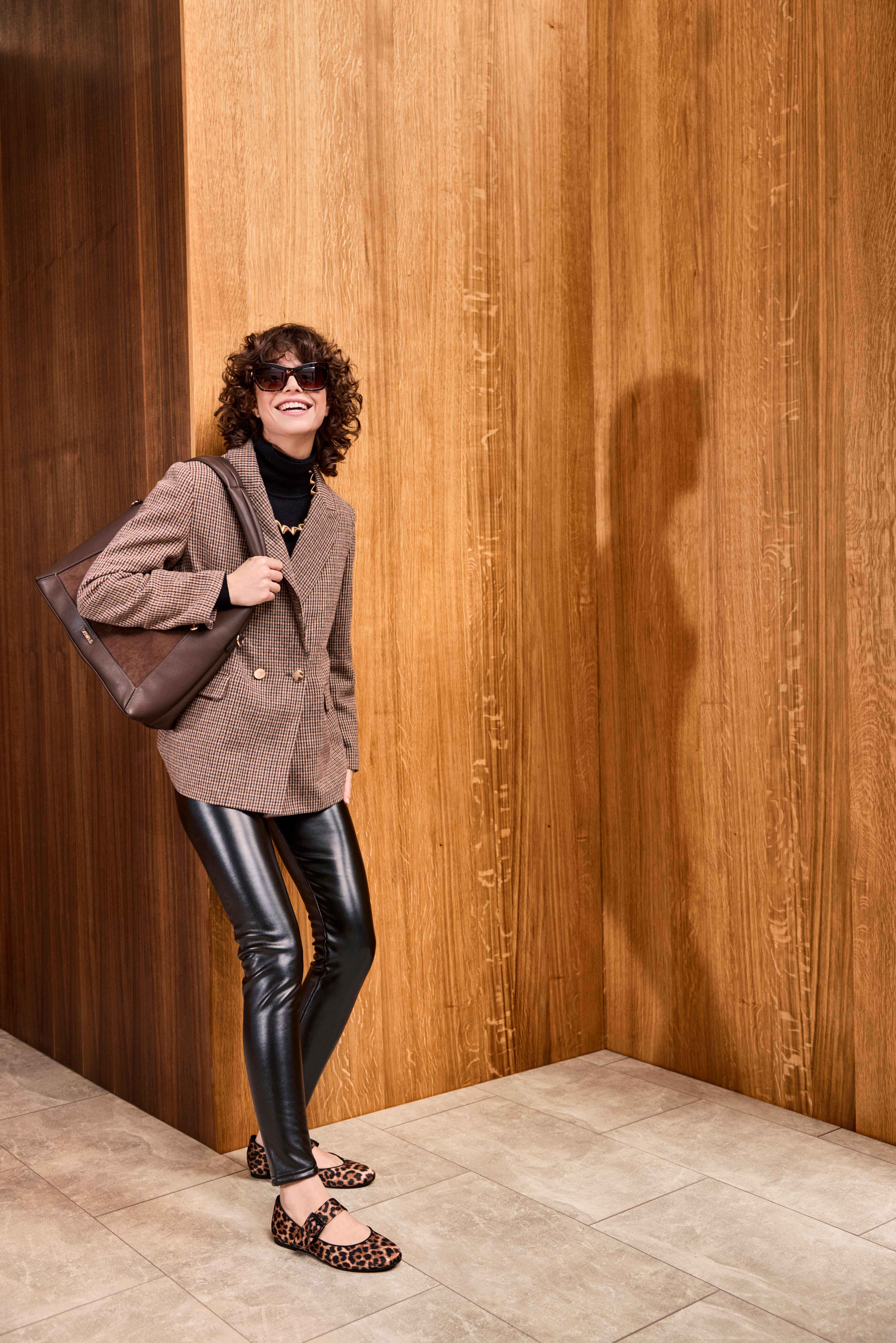 Frau im Office-Look mit Karoblazer, Lederleggings, Loafer mit Leopard-Print undCognac-Tasche, stehend vor Holzwand