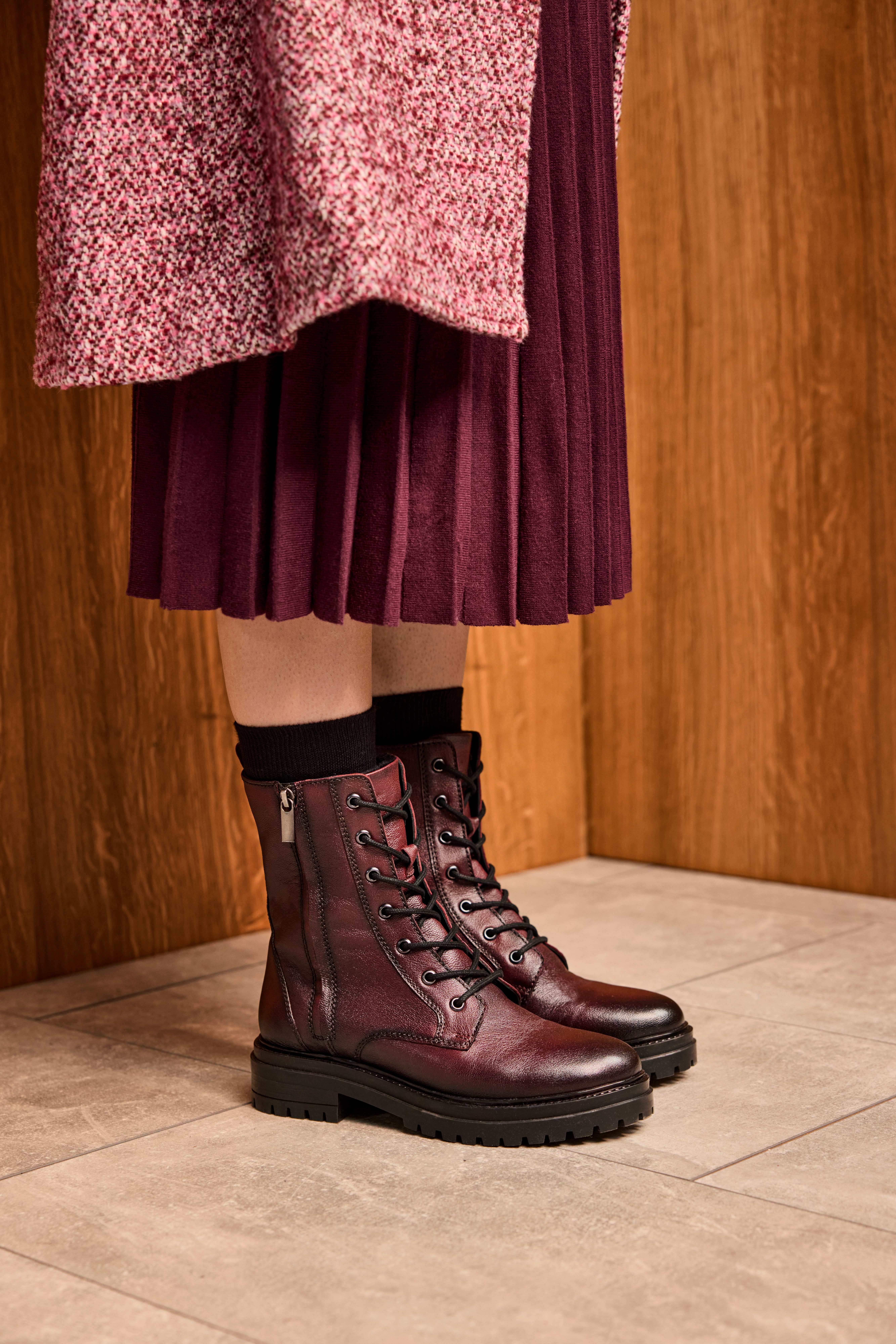 Frau in burgunderrotem Kleid, lehnt an Wand, trägt tiefrote Stiefel aus Leder