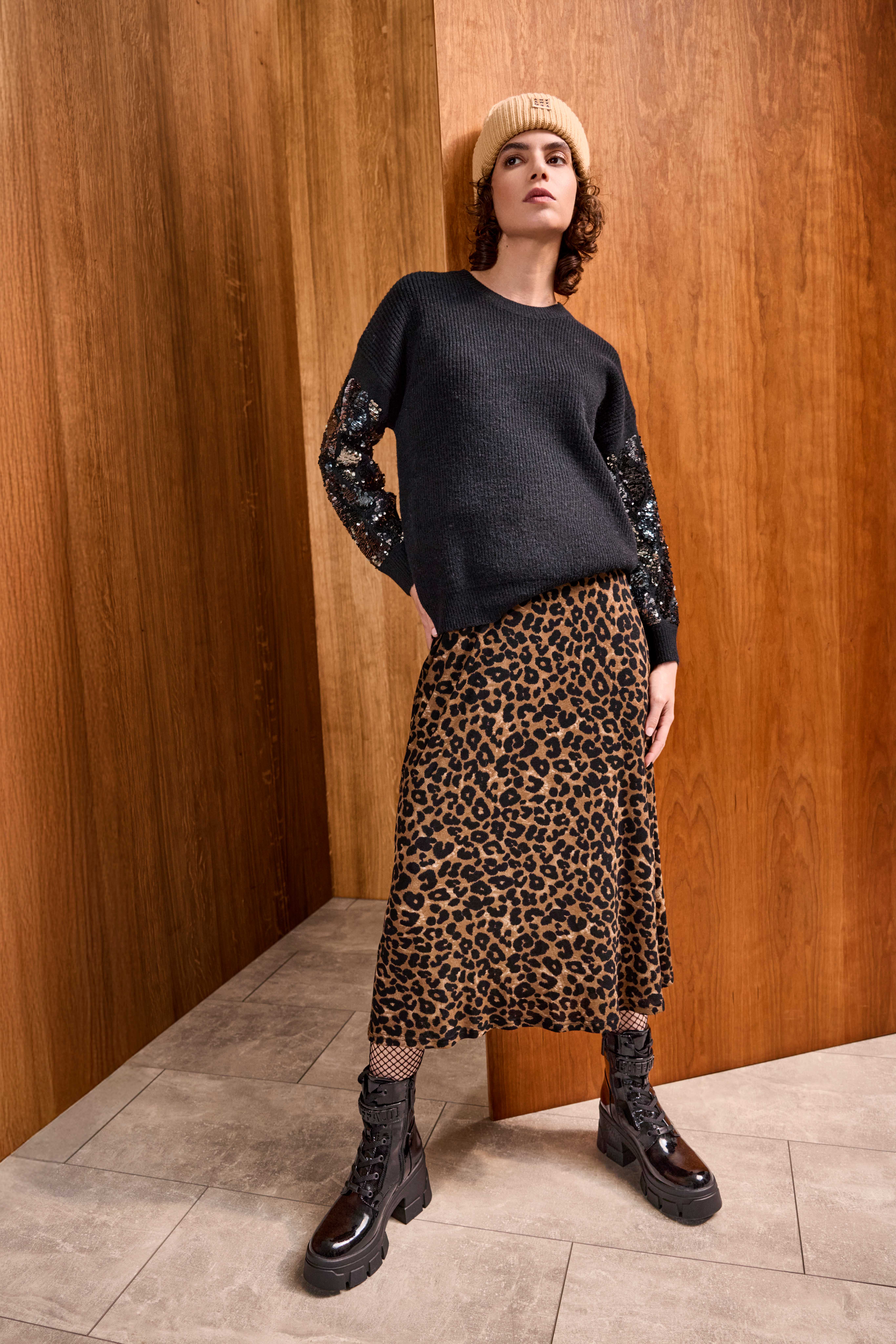 Frau lehnt an Wand, trägt schwarzen Pullover mit Leoparden-Skirt und Beanie