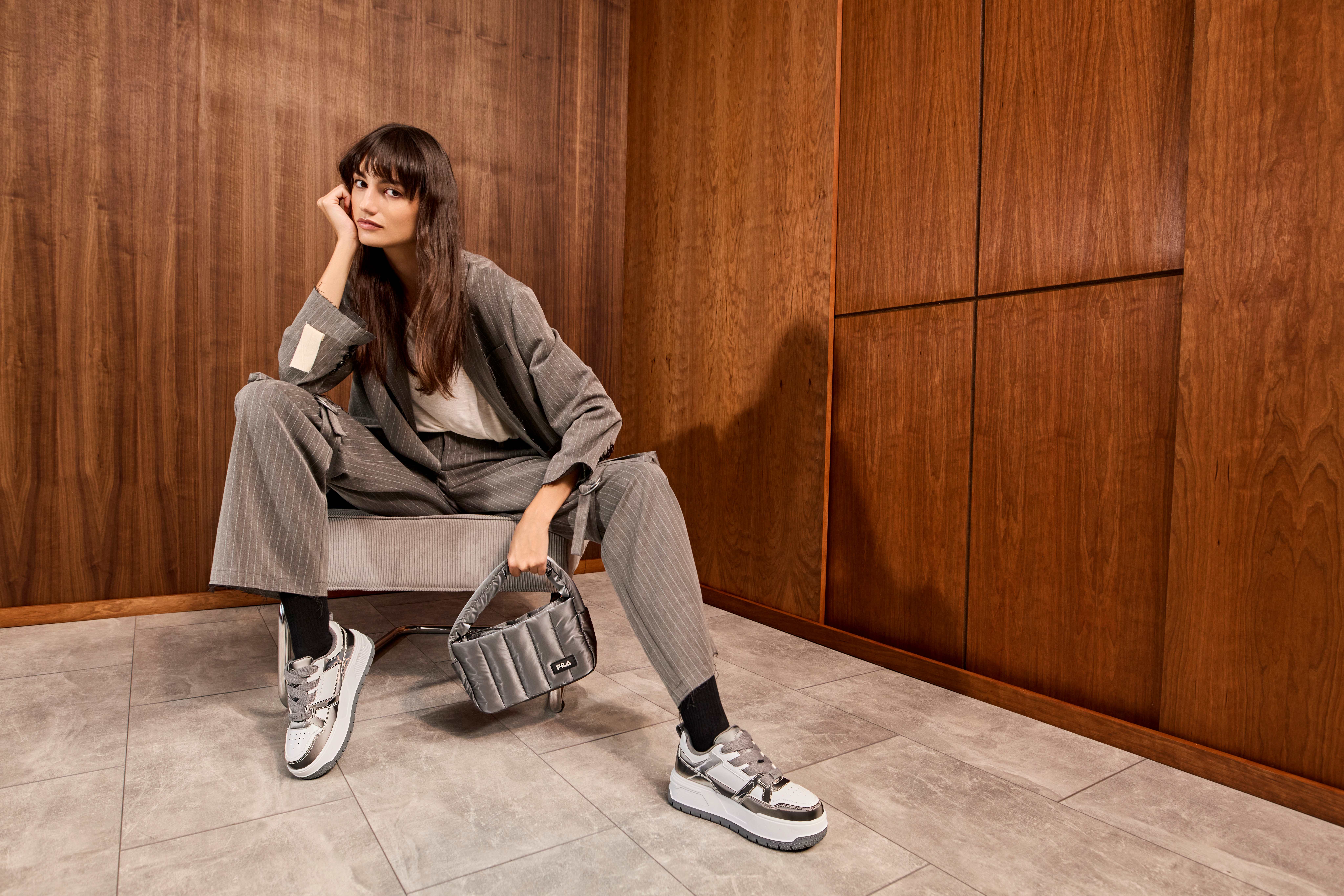Frau in grauem Business-Outfit, sitzt lässig in Sessel, trägt Handtasche und weiße Sneaker