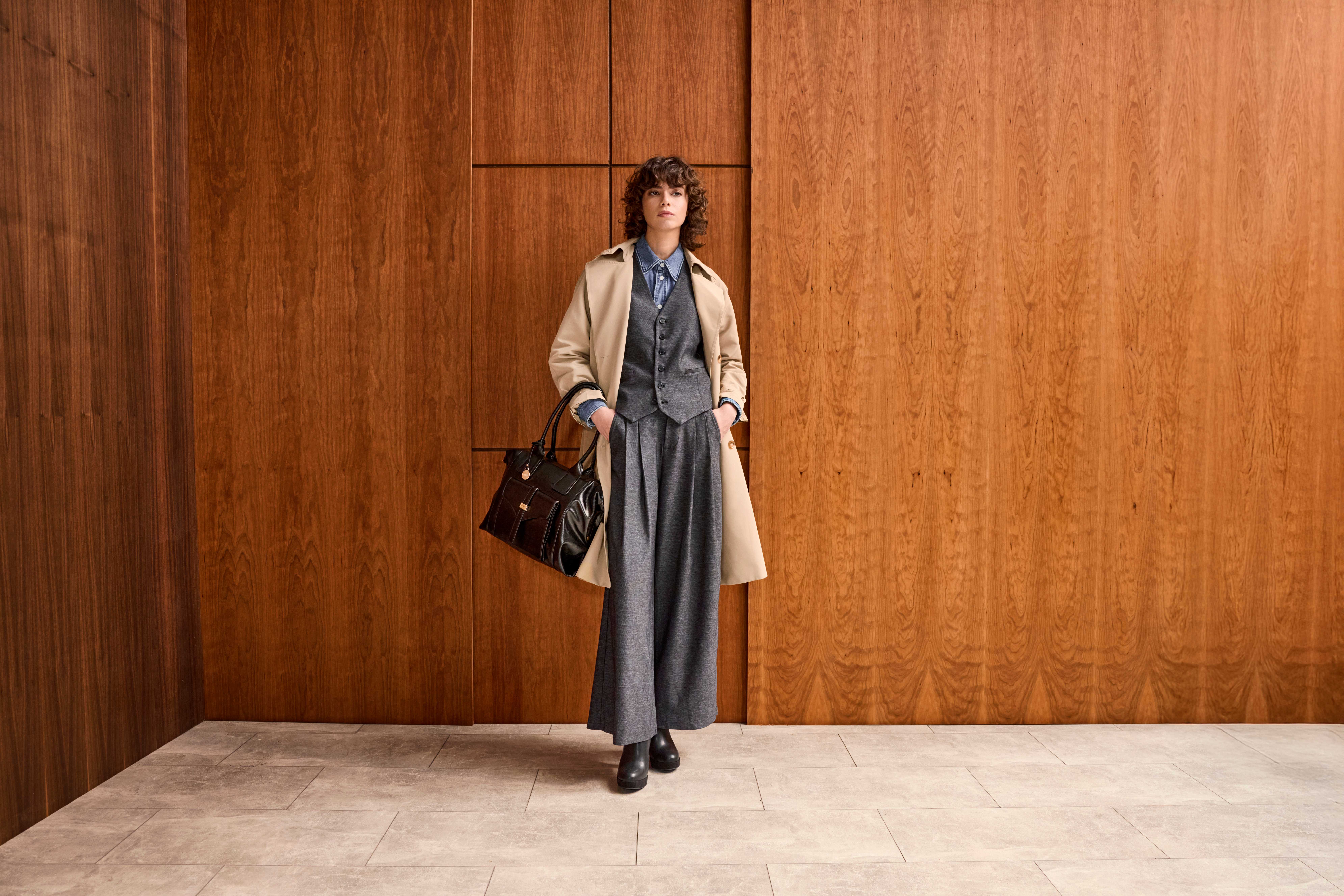Frau in lässigem Office Siren-Look, mit braunem Trenchcoat, stehend vor Holzwand
