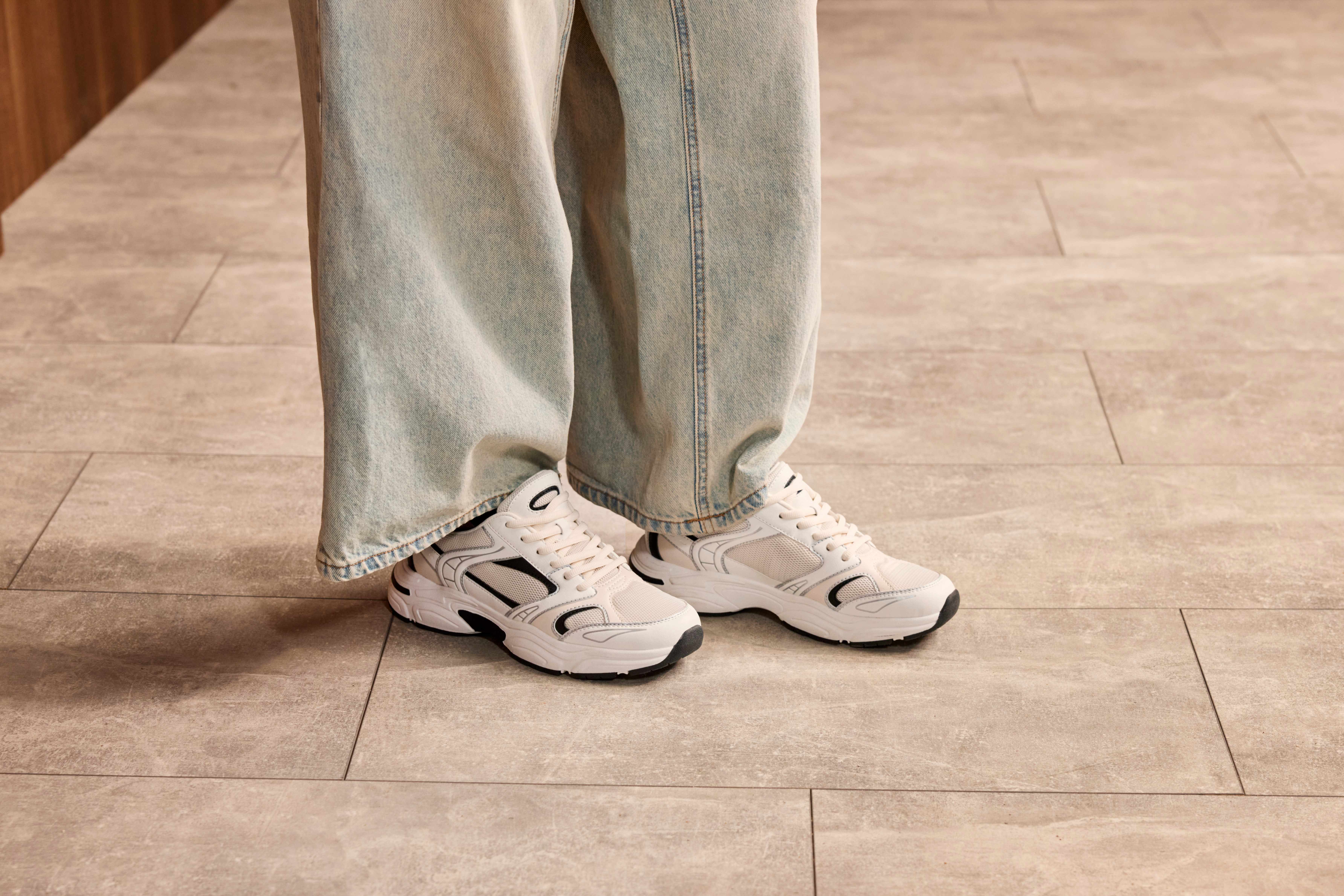 Person trägt weiße Stoffschuhe und ausgewaschene Jeanshose, steht auf Fliesenboden