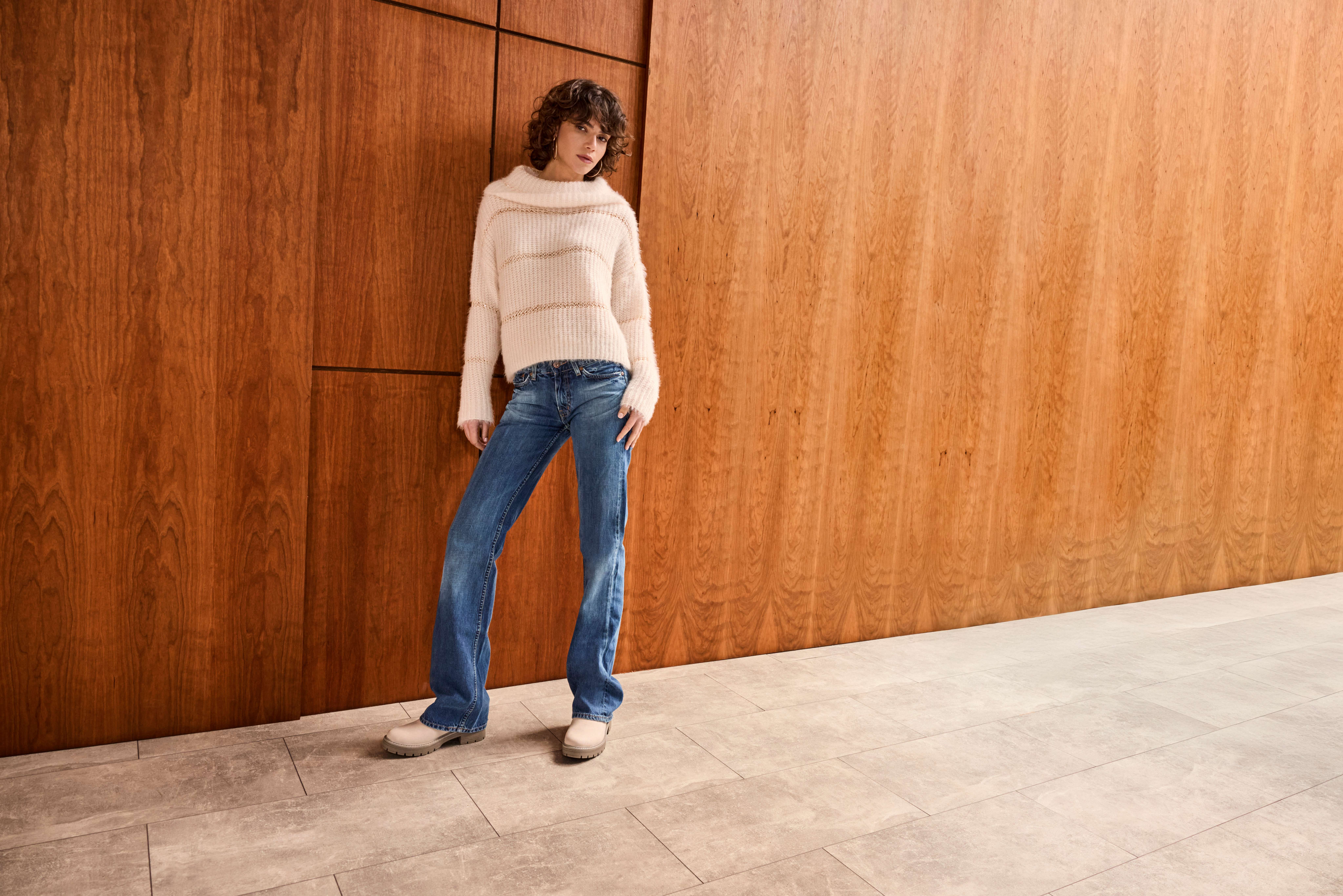 Frau posiert in weißem Sweater und blauer Jeans vor einer Holzwand