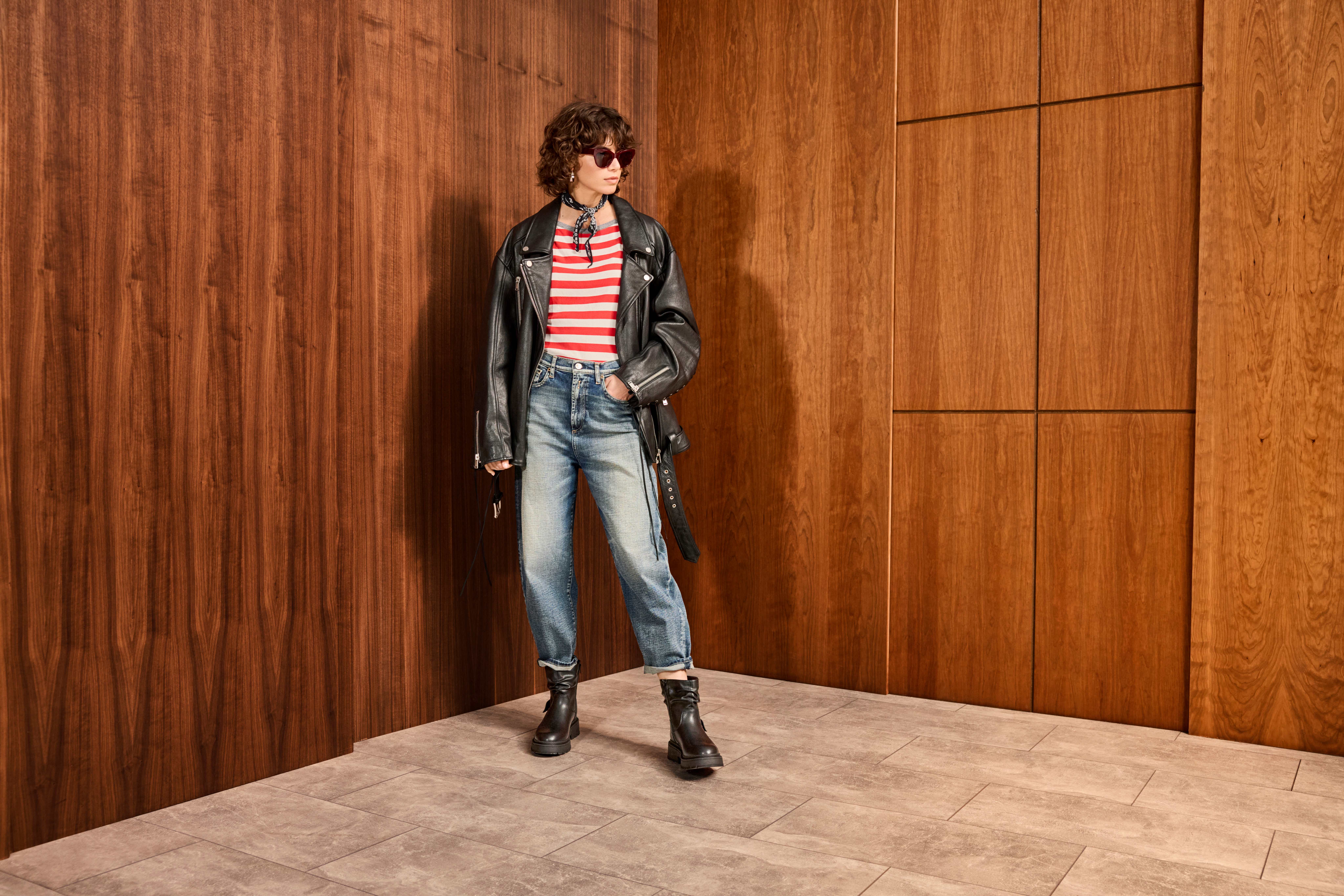 Frau in schwarzem Mantel, mit weiß-rot gestreiftem Shirt und Jeanshose, steht lässig vor Holzwand