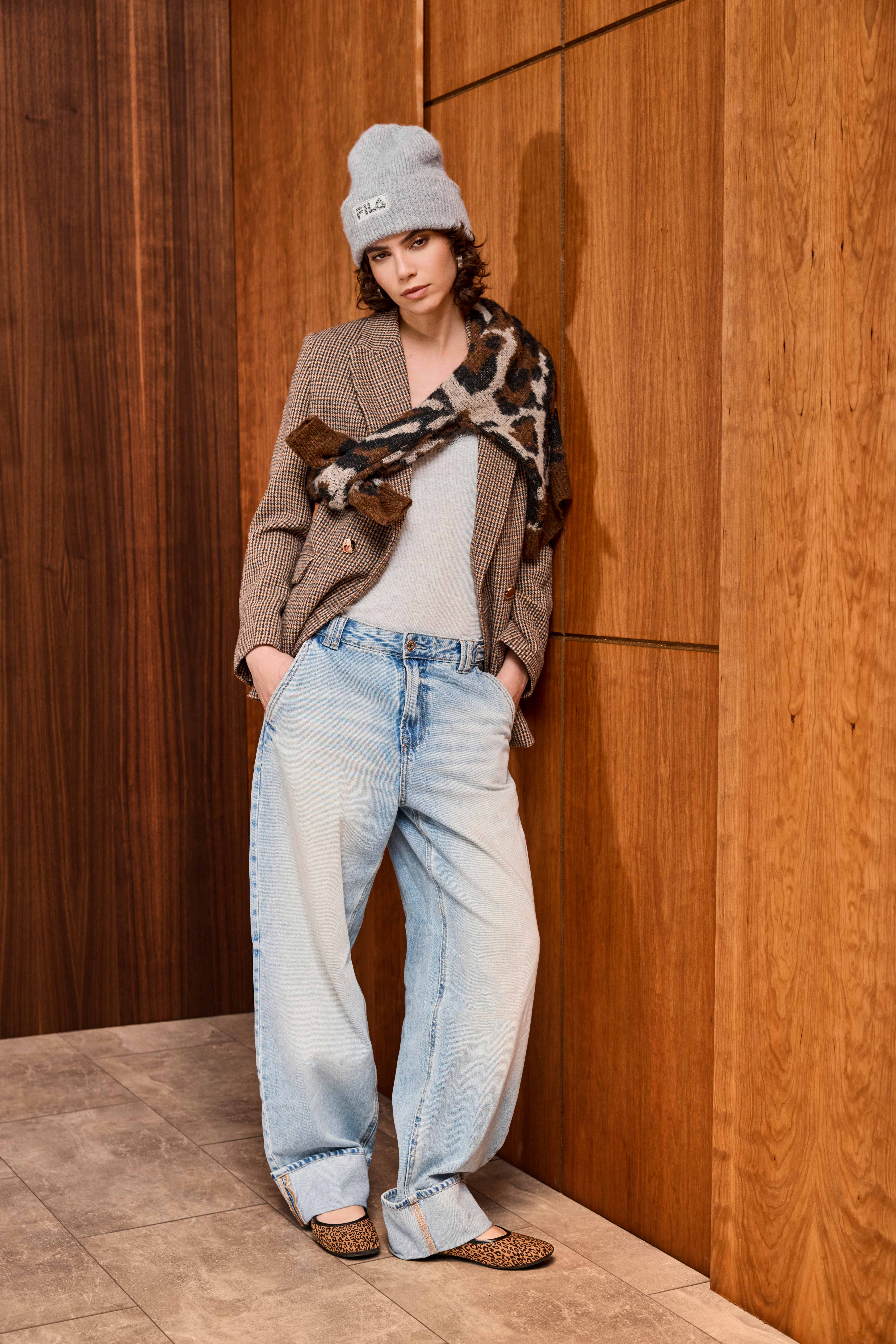 Eine Frau trägt einen karierten Blazer, ein graues Shirt, eine weite Jeans, eine graue Mütze und Leo-Ballerinas
