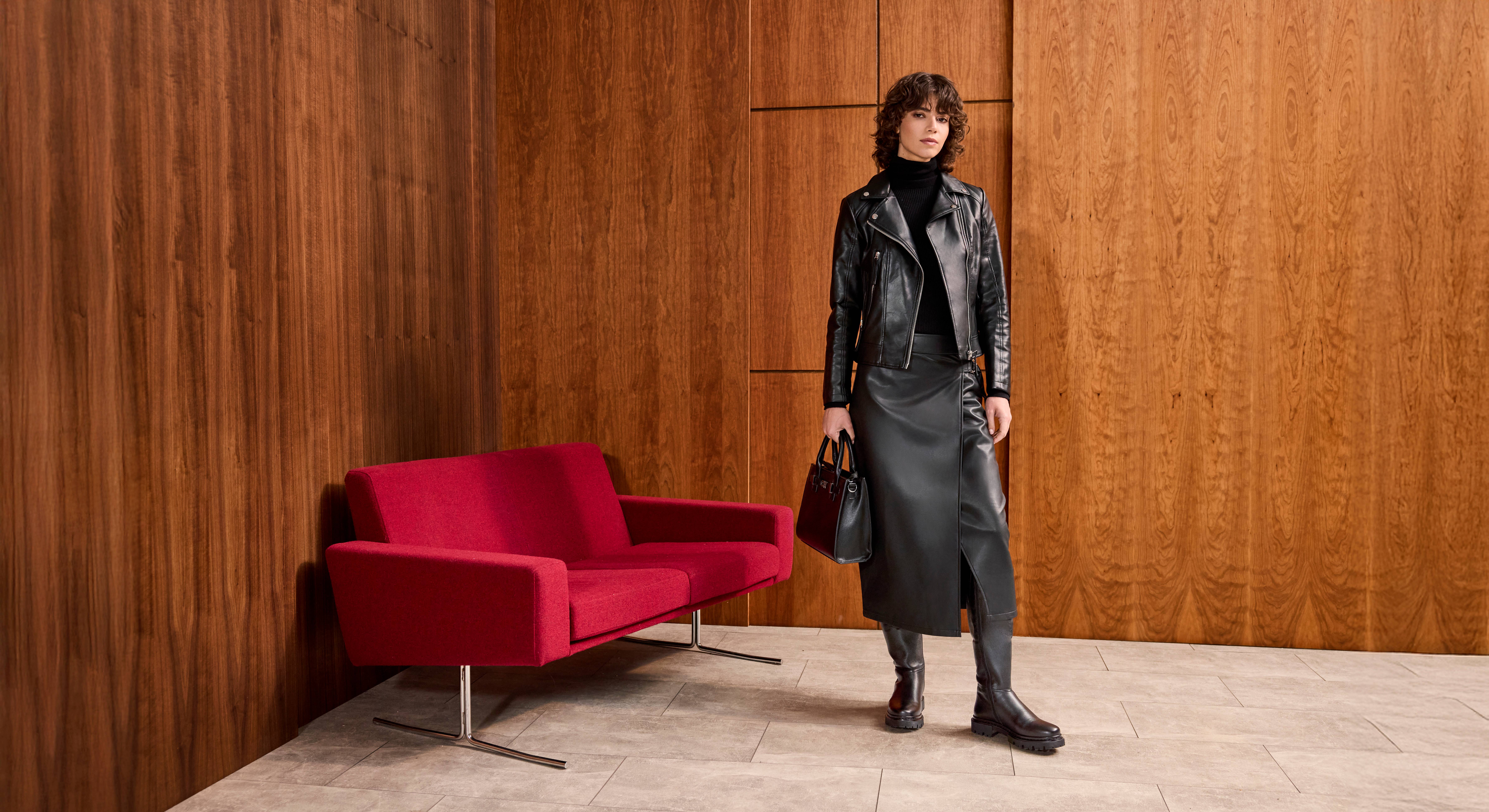 Frau in schwarzem Leder-Outfit, mit passenden Boots und Handtasche, steht neben rotem Sofa