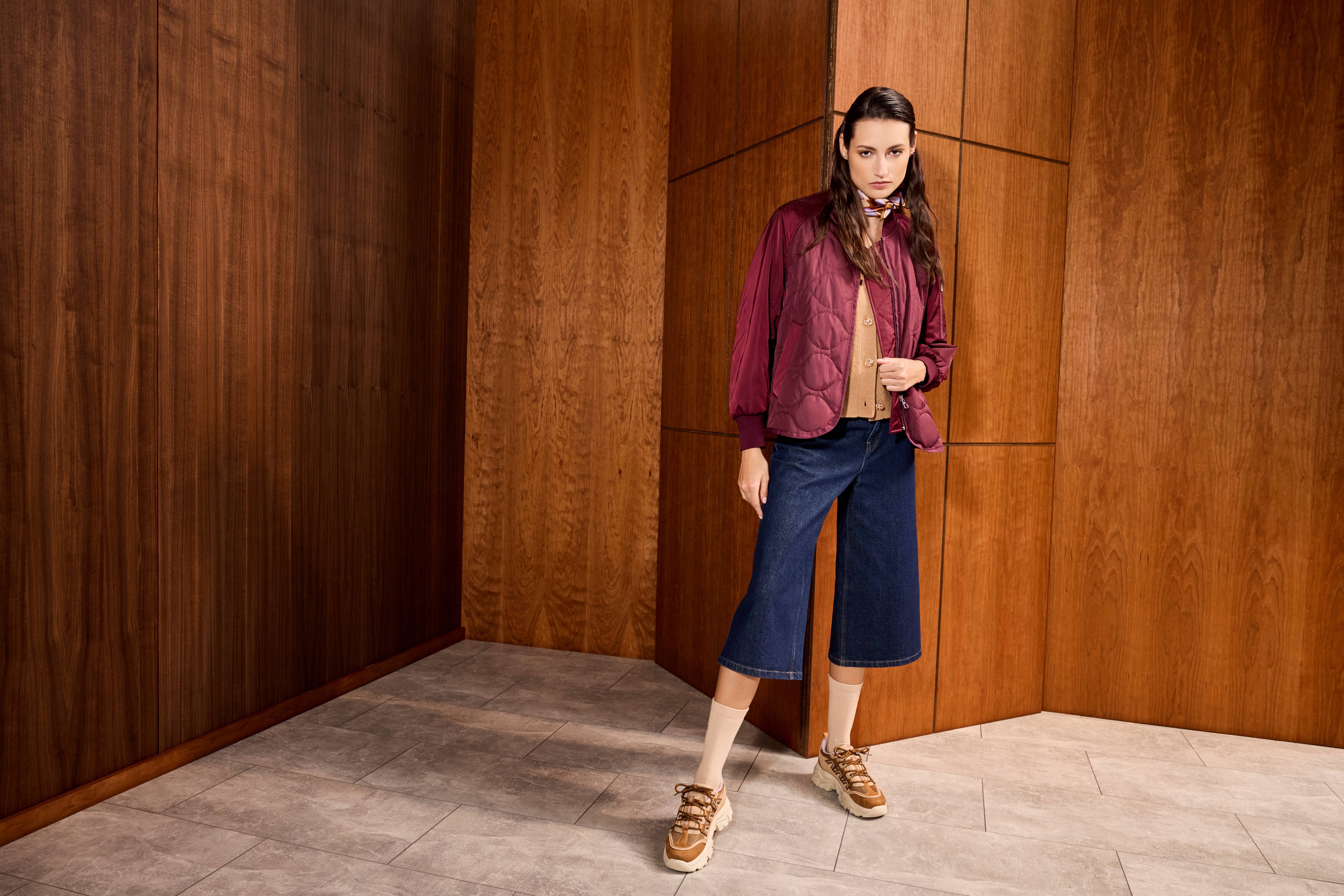 Frau in roter Jacke, blauer Jeanshose und braunen Dad Sneakern, stehend vor Holzwand