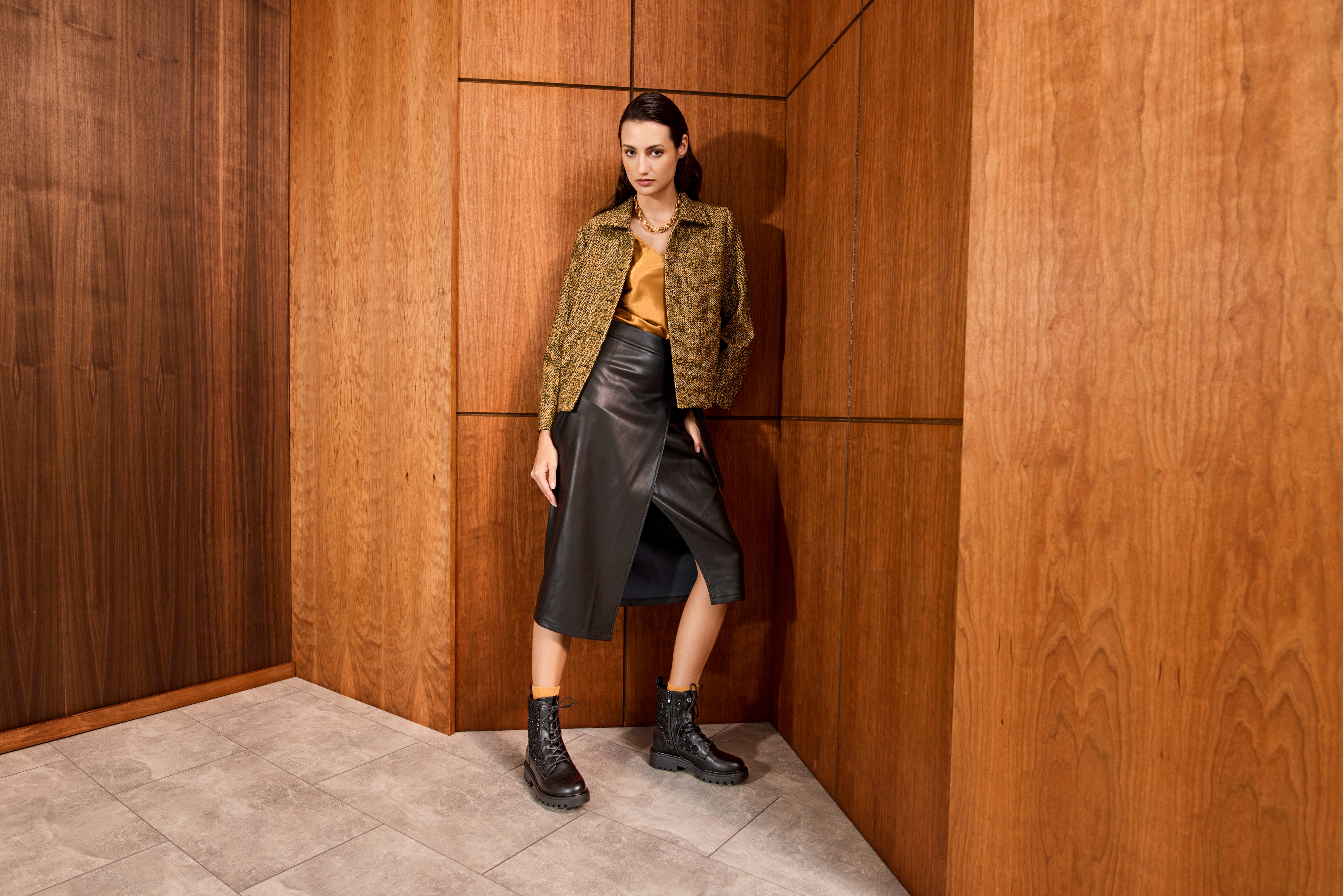 Frau steht vor brauner Holzwand, trägt schwarzen Leder-Skirt in Kombination mit schwarzen Boots