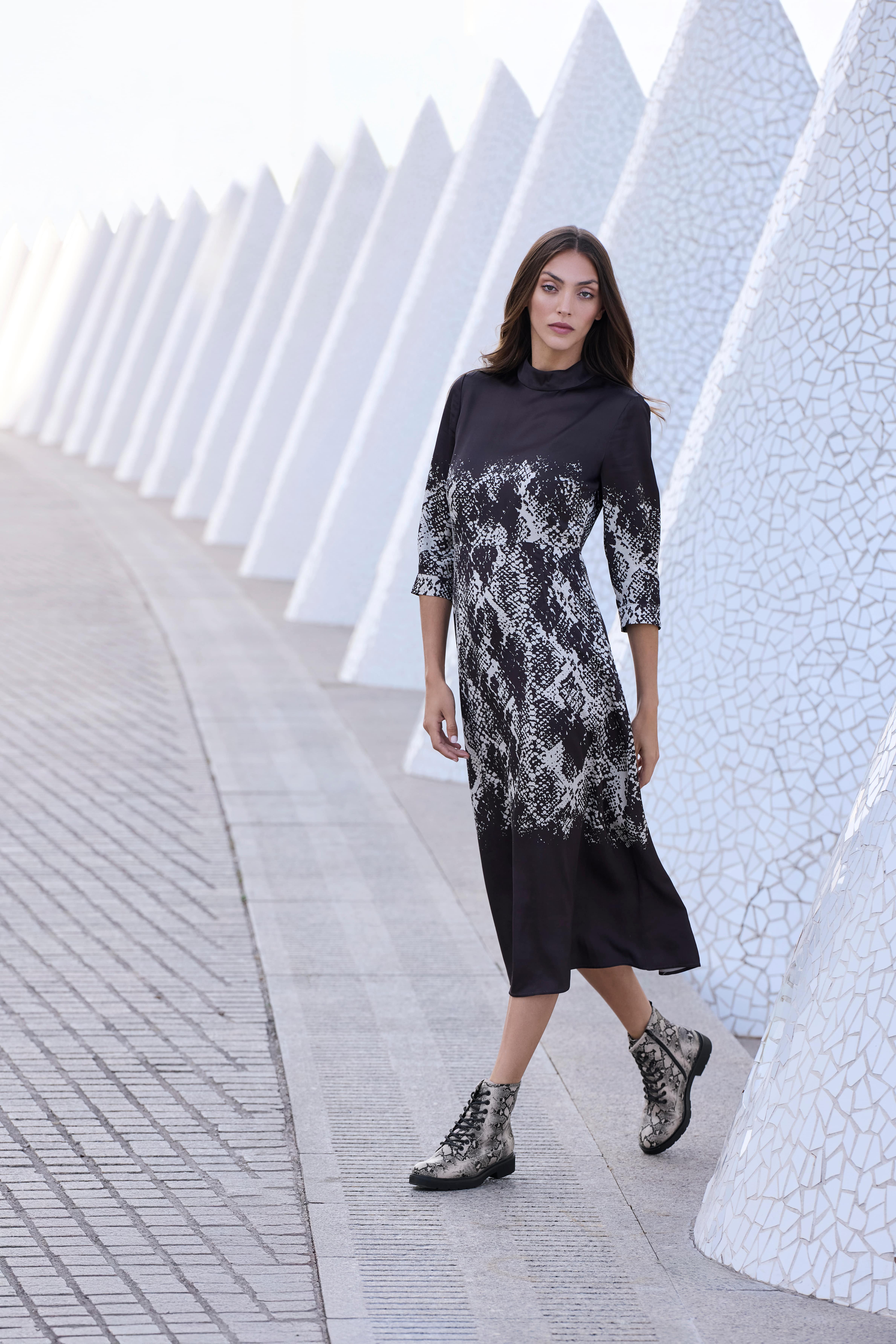 Eine Frau trägt ein schwarzes Midikleid mit Schlangenprint-Details und dazu passende geschnürte Stiefeletten
