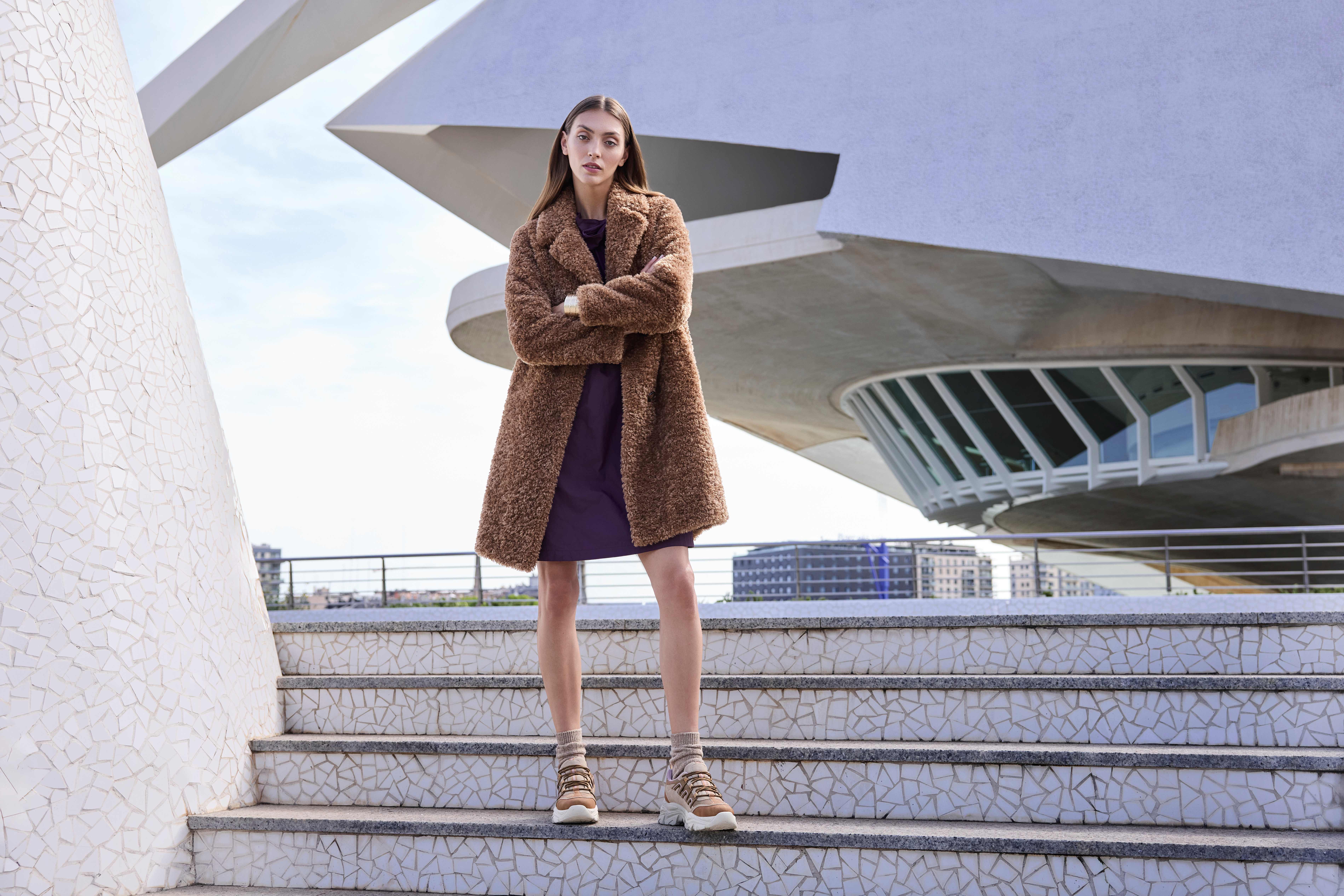Eine Frau steht mit verschränkten Armen auf einer Treppe und trägt einen braunen Teddy-Mantel, ein lila Kleid und beige Sneakers mit Socken