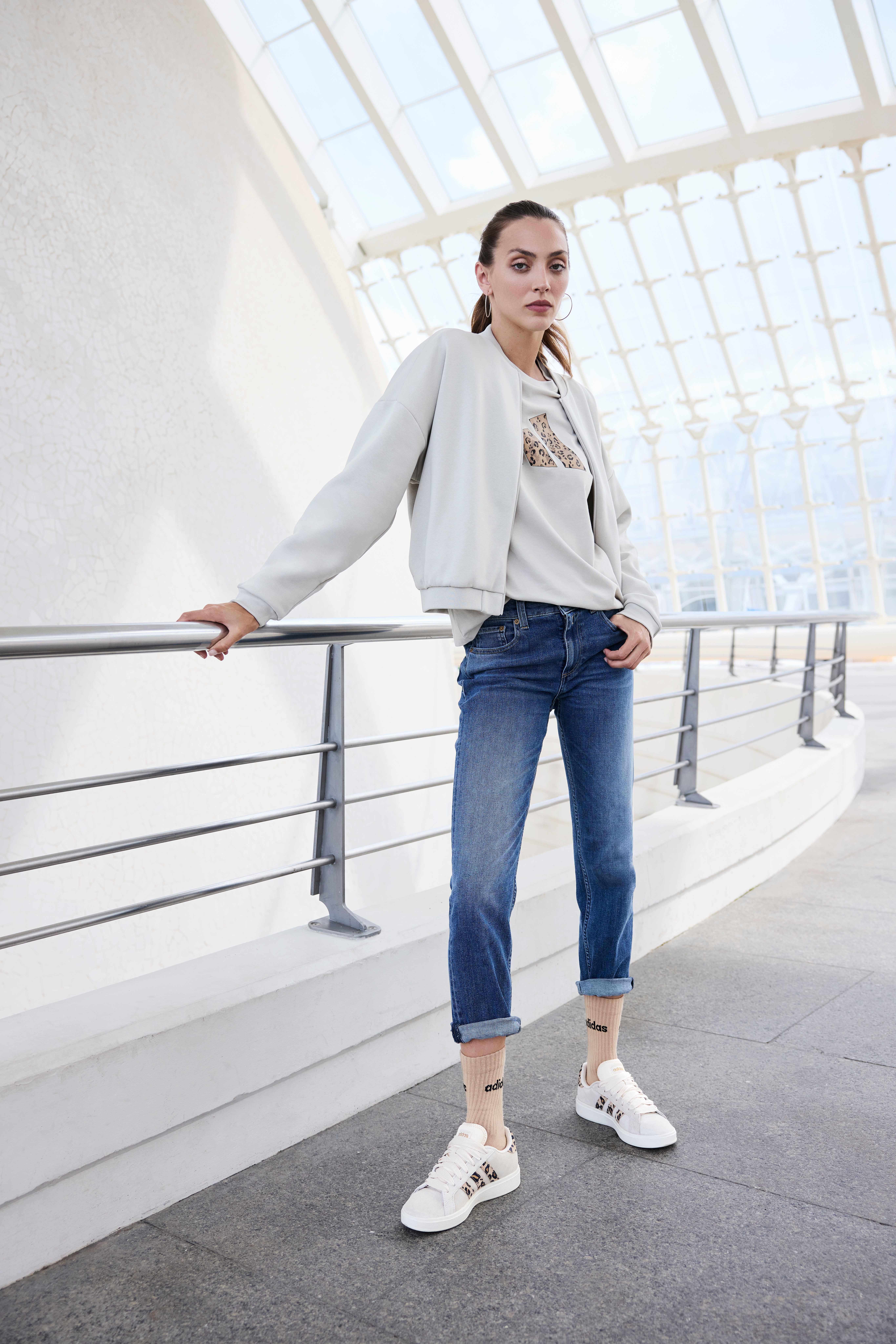 Eine Frau trägt eine hellgraue Bomberjacke, ein bedrucktes Shirt, eine blaue Jeans und weiße Sneakers
