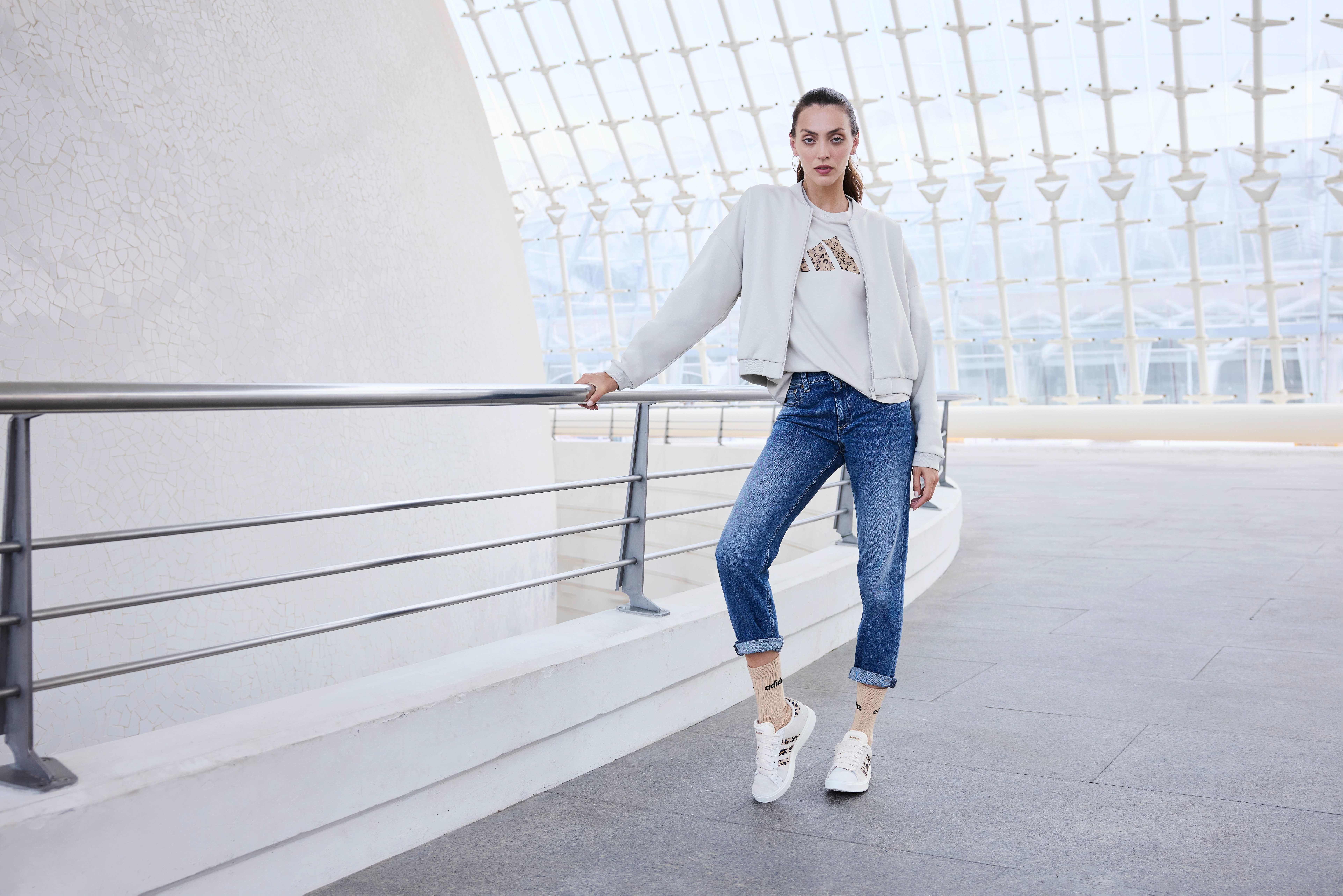 Frau mit weißer Strickjacke und Adidas-Shirt, trägt lange Jeanshose und weiße Sneaker, lehnt an Metallgeländer