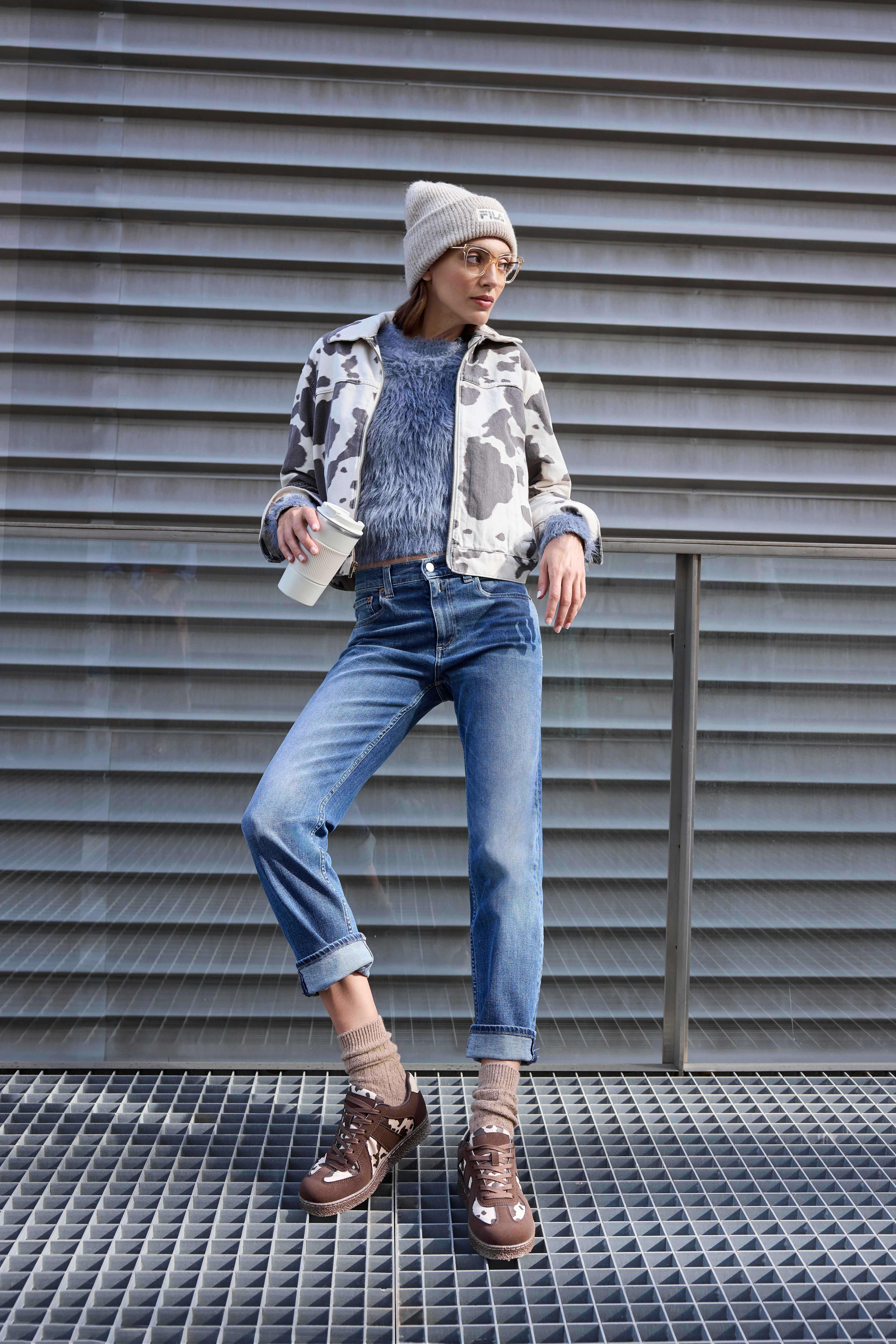 Eine Frau trägt eine Jeans mit hochgekrempelten Beinen, einen flauschigen blauen Pullover unter einer Kuhflecken-Jacke, braune Sneakers und eine graue Beanie