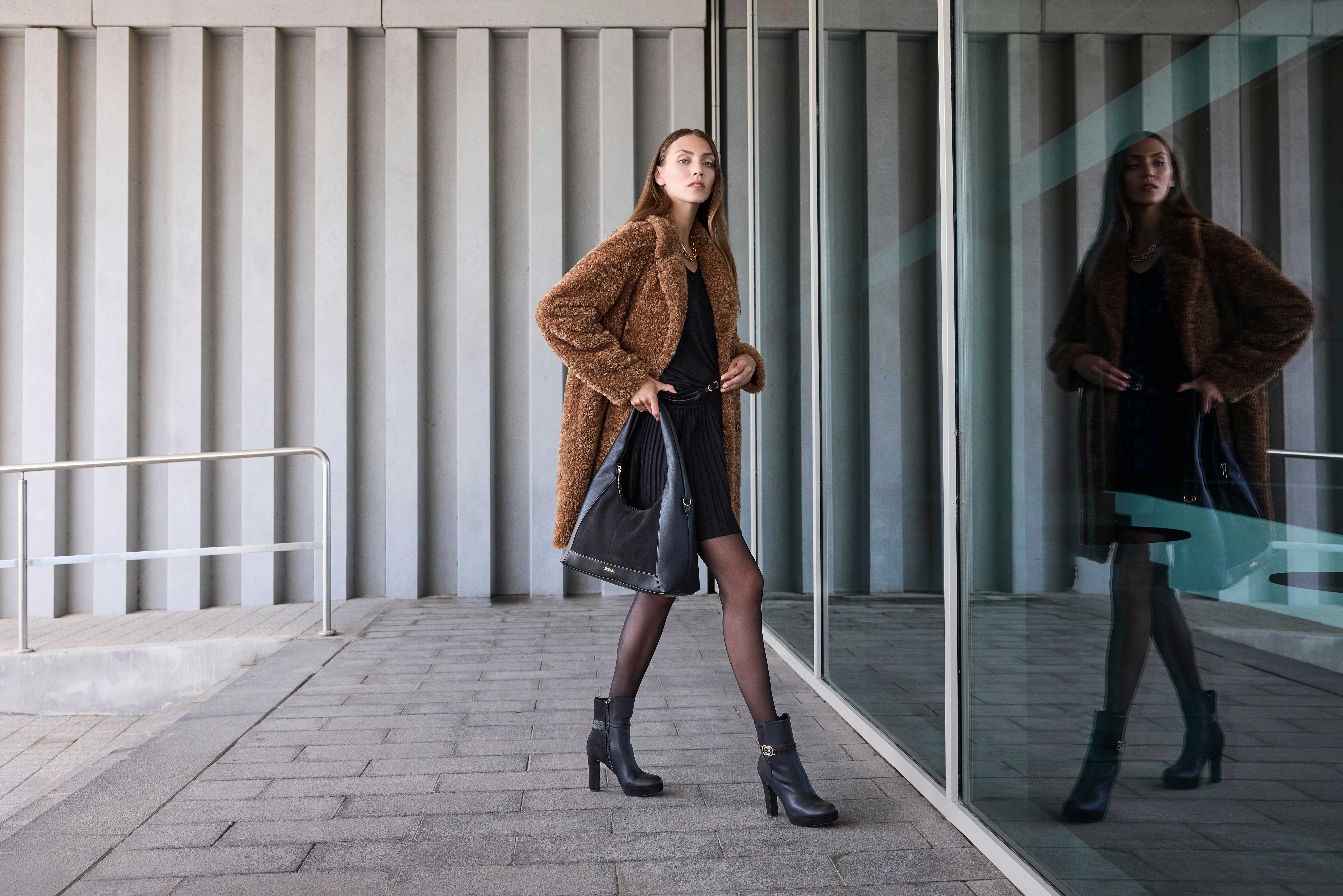Eine Frau trägt ein schwarzes Kleid mit kurzem Rock, kombiniert mit einem braunen Teddy-Mantel, schwarzen Stiefeletten und einer großen schwarzen Tasche