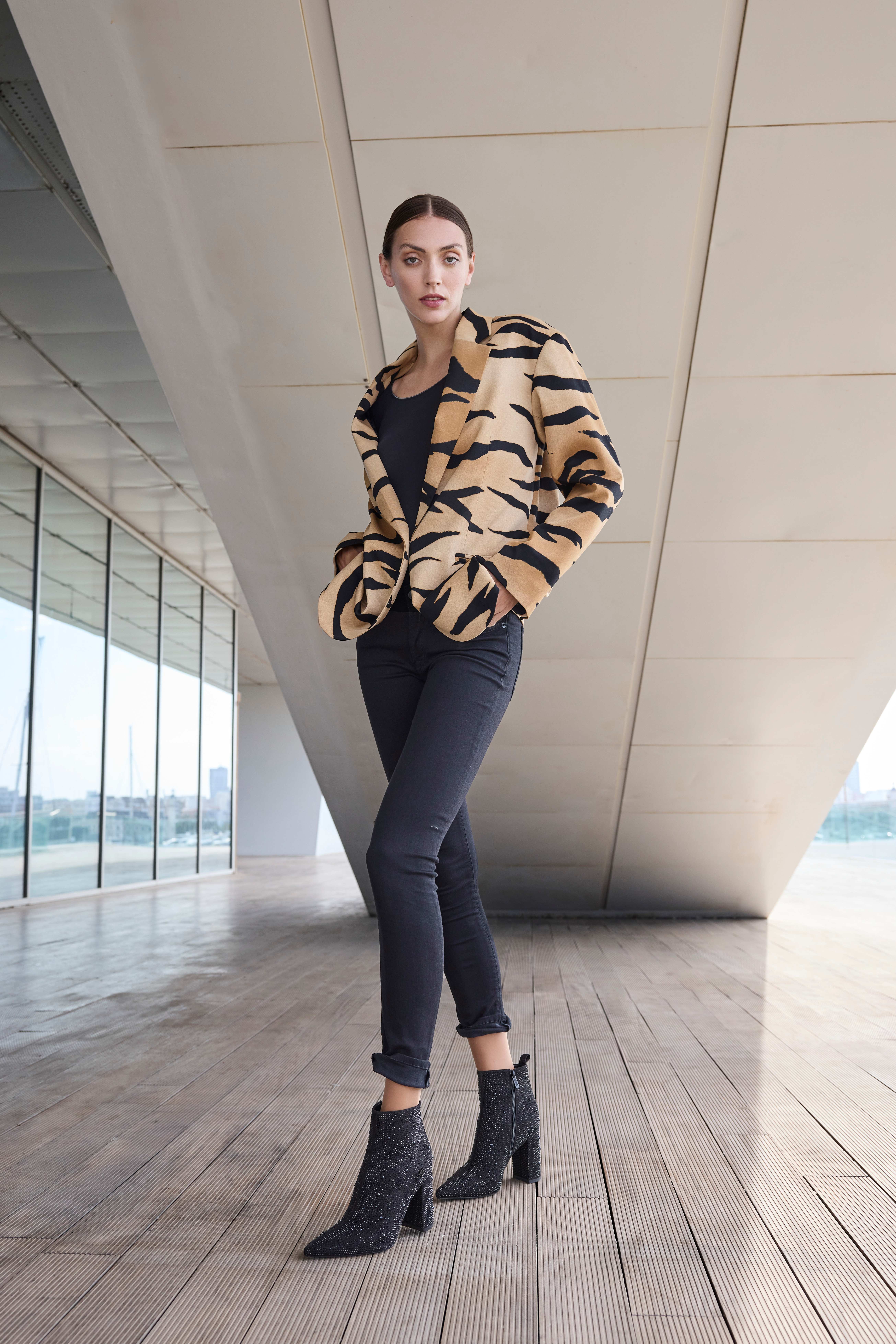 Eine Frau trägt eine enge schwarze Hose mit einem schwarzen Top und einem auffälligen Tigerstreifen-Blazer sowie schwarzen Absatzstiefeletten