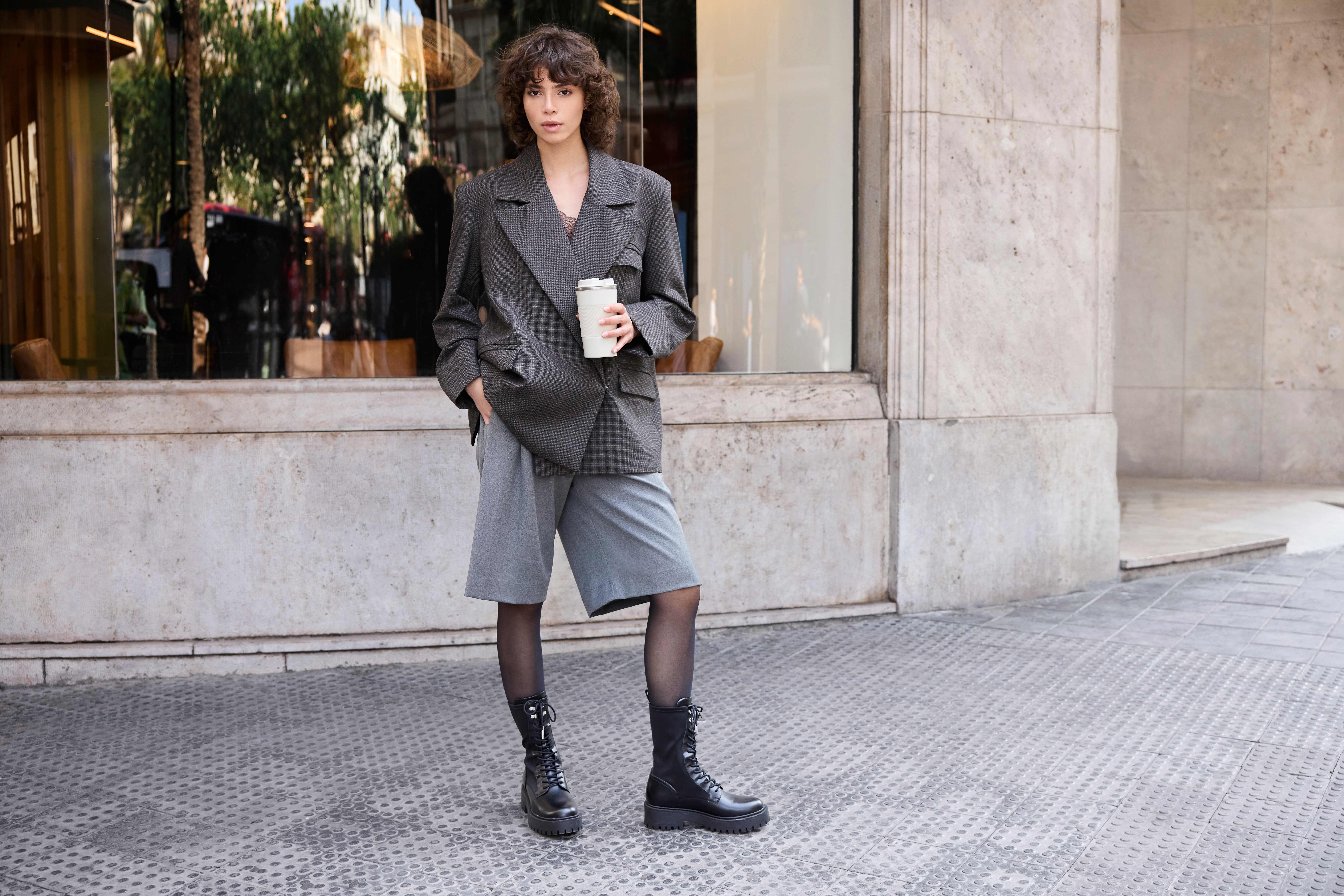Frau mit lockigem Haar in grauem Blazer mit lässiger Short und schwarzen Boots