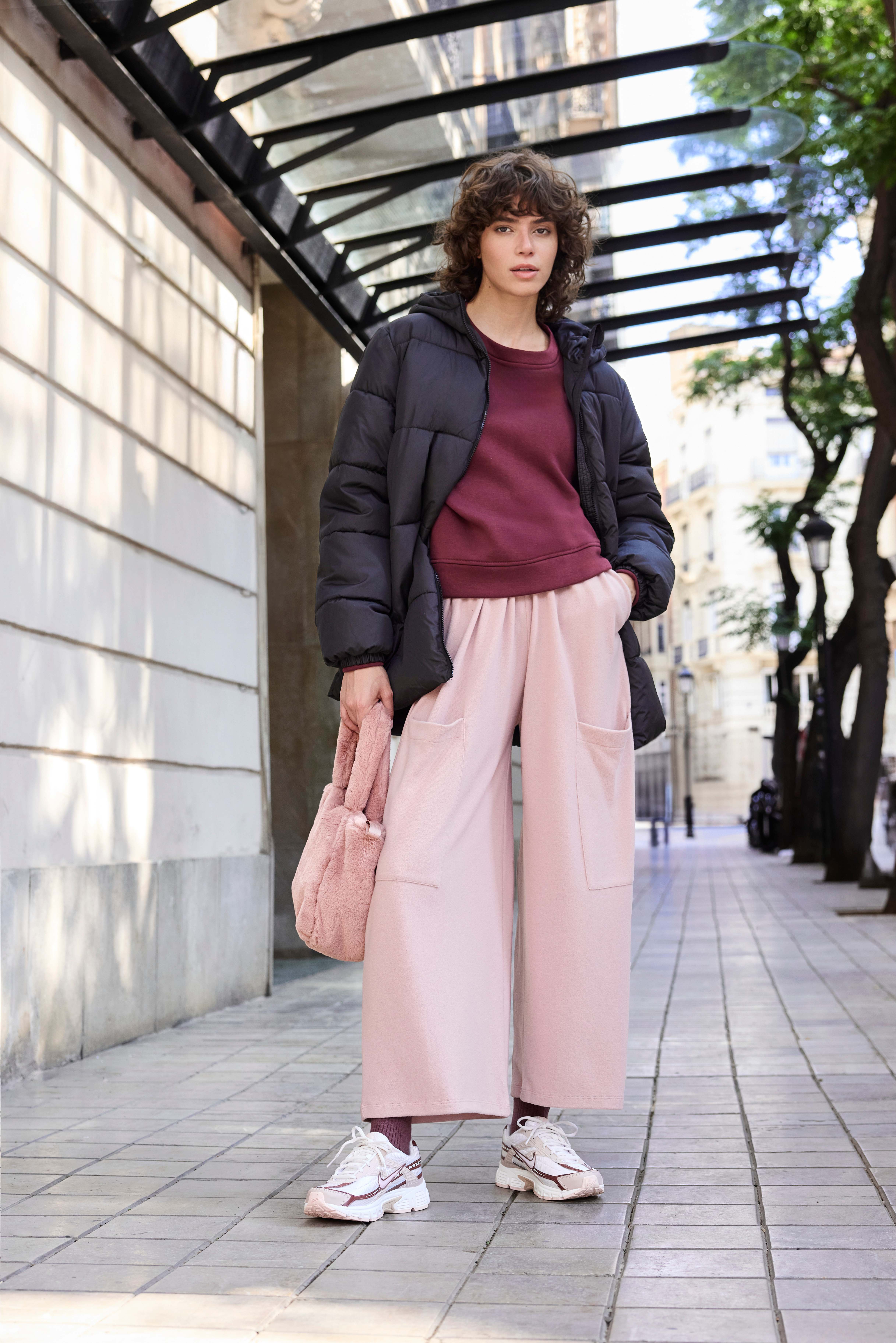 Eine Frau trägt eine schwarze Steppjacke, ein bordeauxfarbenes Sweatshirt, eine rosafarbene Hose und weiße Sneakers