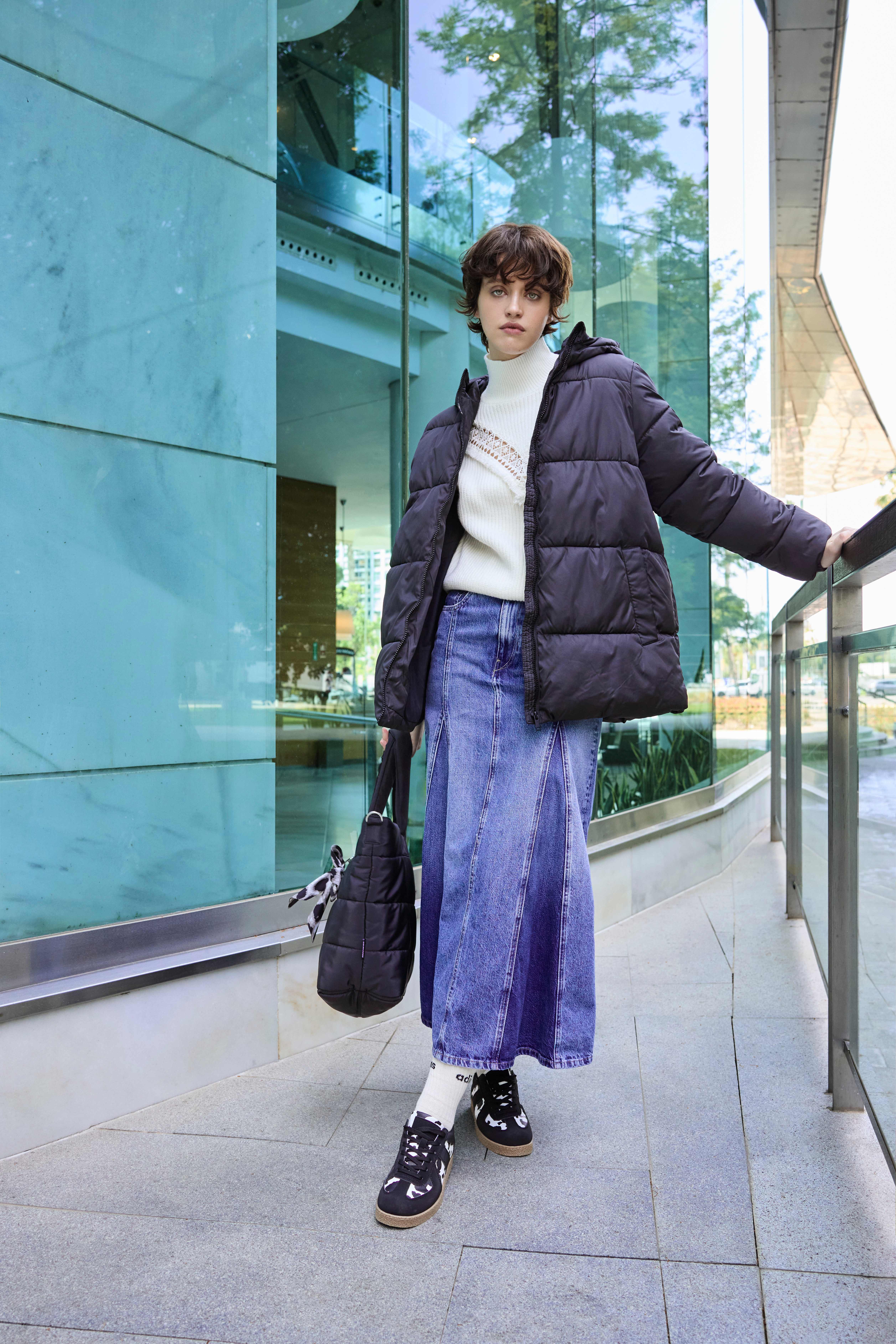 Frau in lila Denim-Maxirock, kuscheligem Strickpulli und Pufferjacke mit schwarzen Sneakern