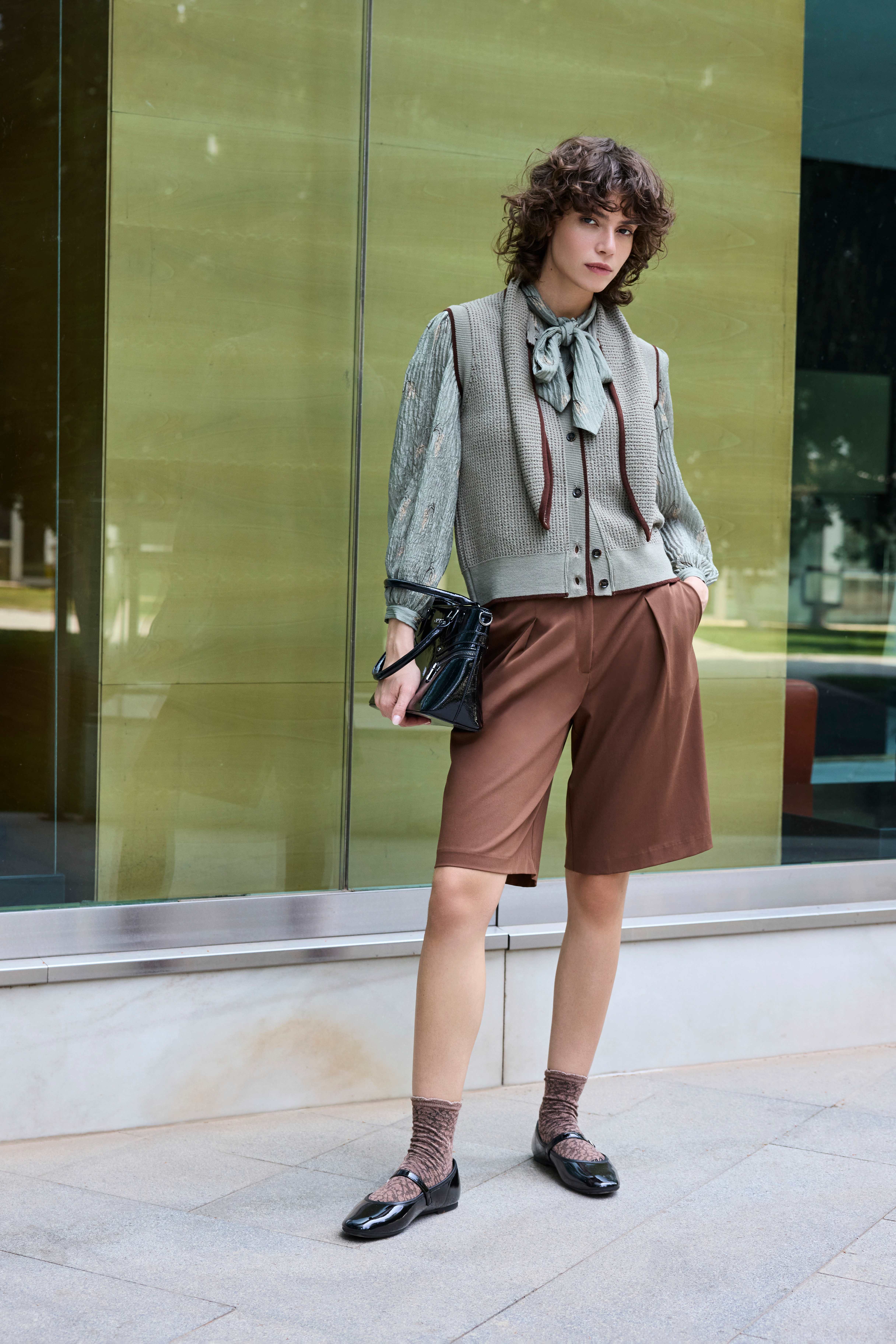Eine Frau steht und trägt eine hellgrüne Bluse, braune Shorts, schwarze Ballerinas und eine schwarze Tasche 