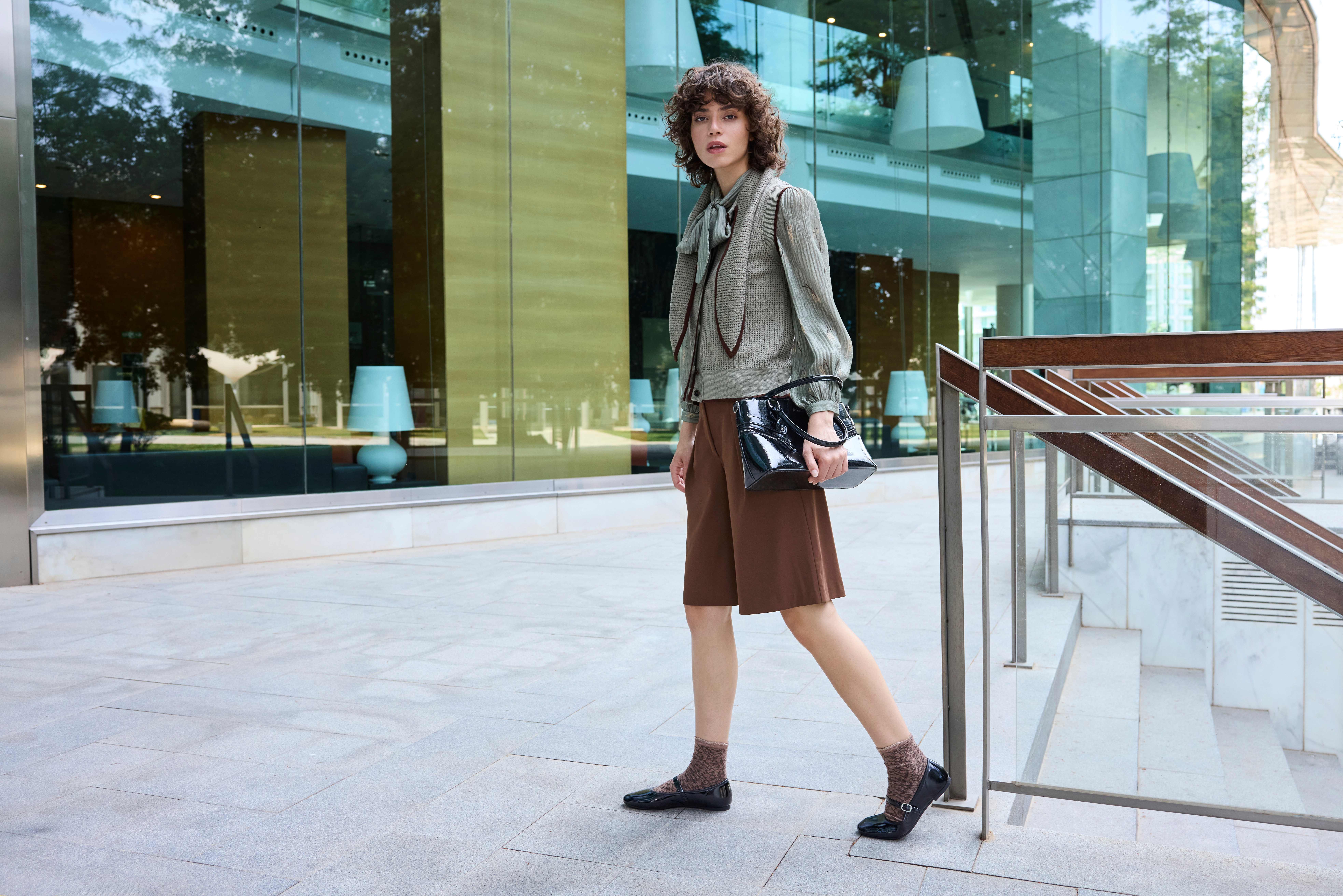 Eine Frau trägt eine hellgrüne Bluse, braune Shorts, schwarze Ballerinas und eine schwarze Tasche 