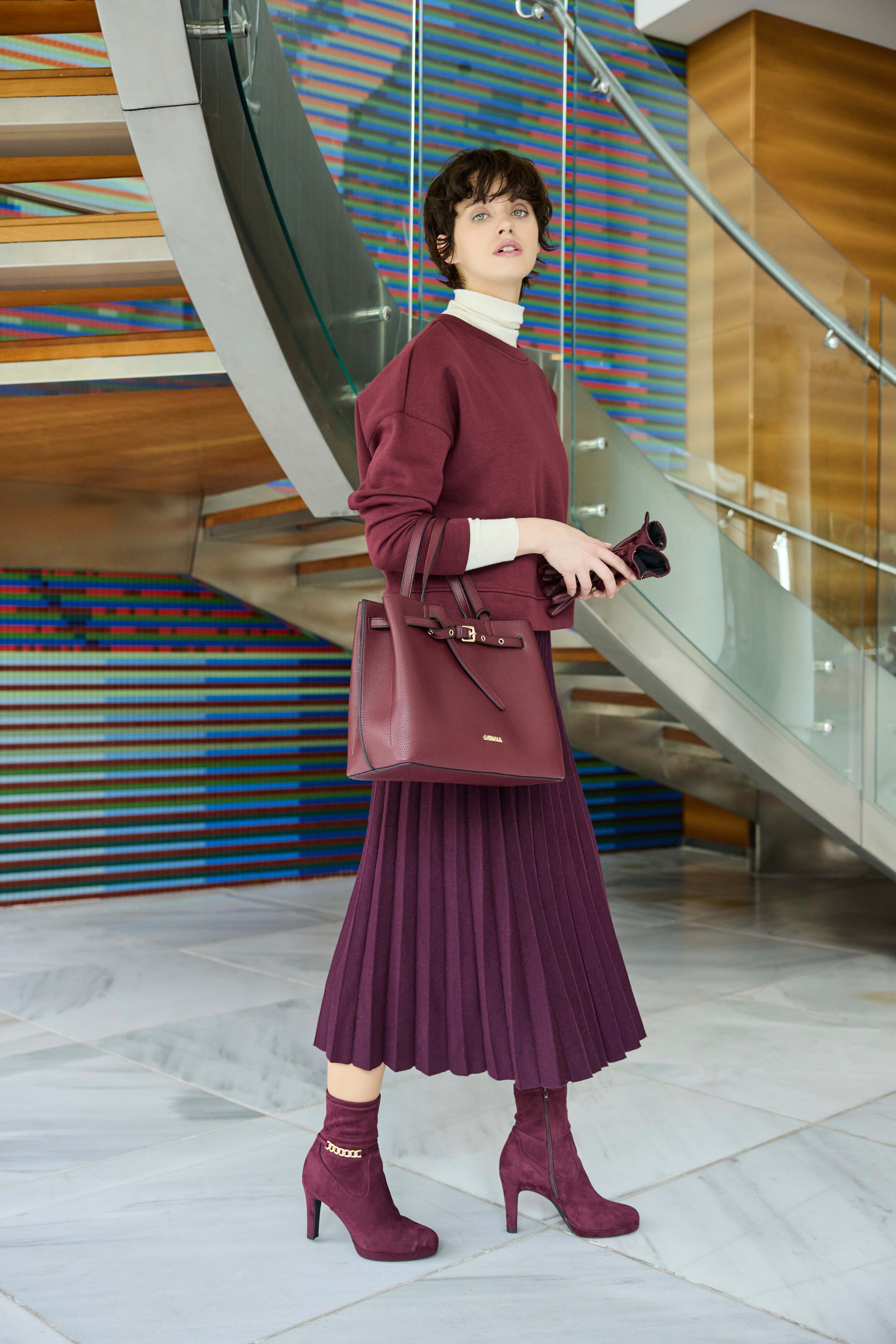Frau in burgunderrotem Pullover mit passendem Faltenrock, Pumps und Handtasche, vor gläserner Wendeltreppe