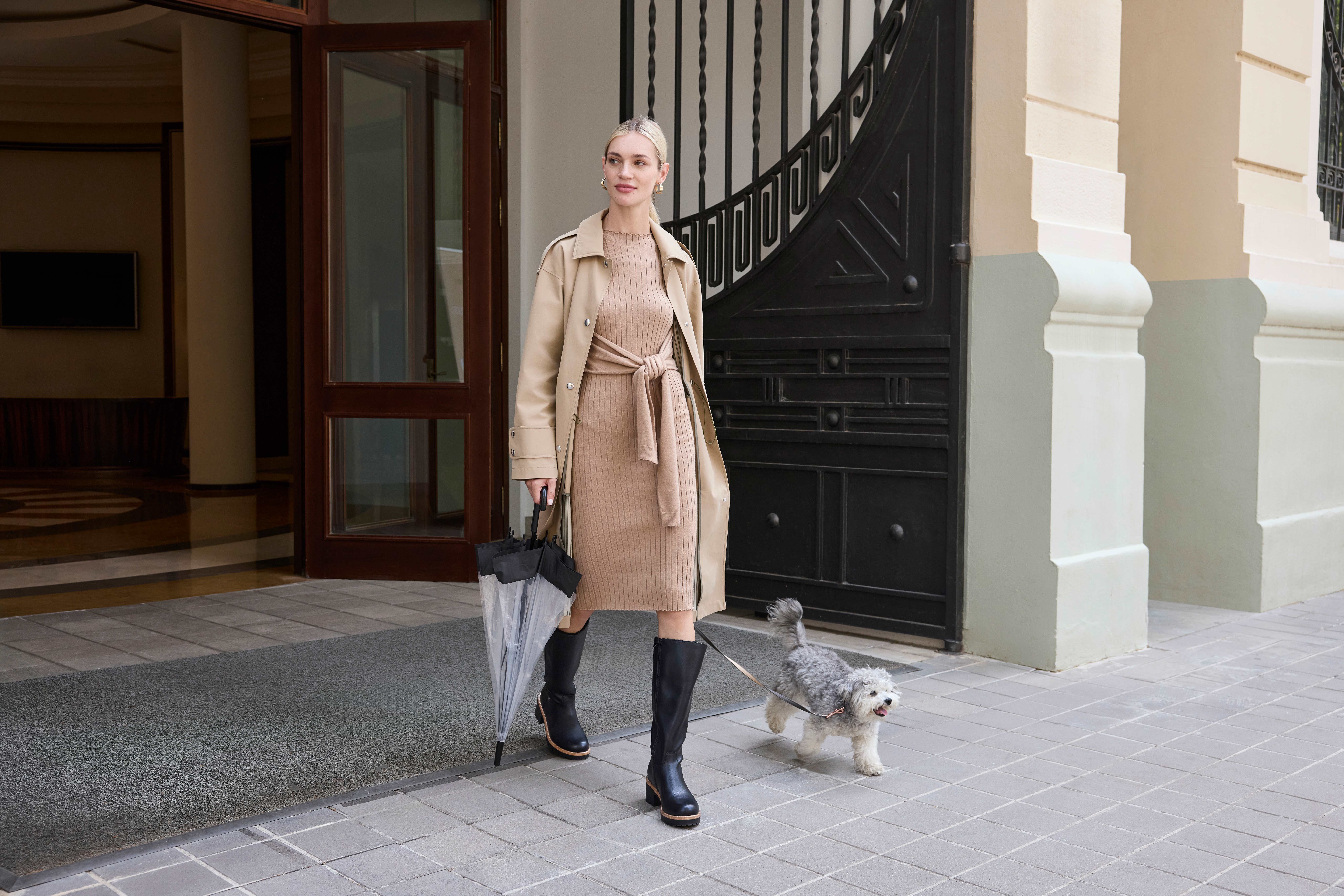 Frau in braunem Trenchcoat und schwarzen Stiefeln geht Gassi mit Hund