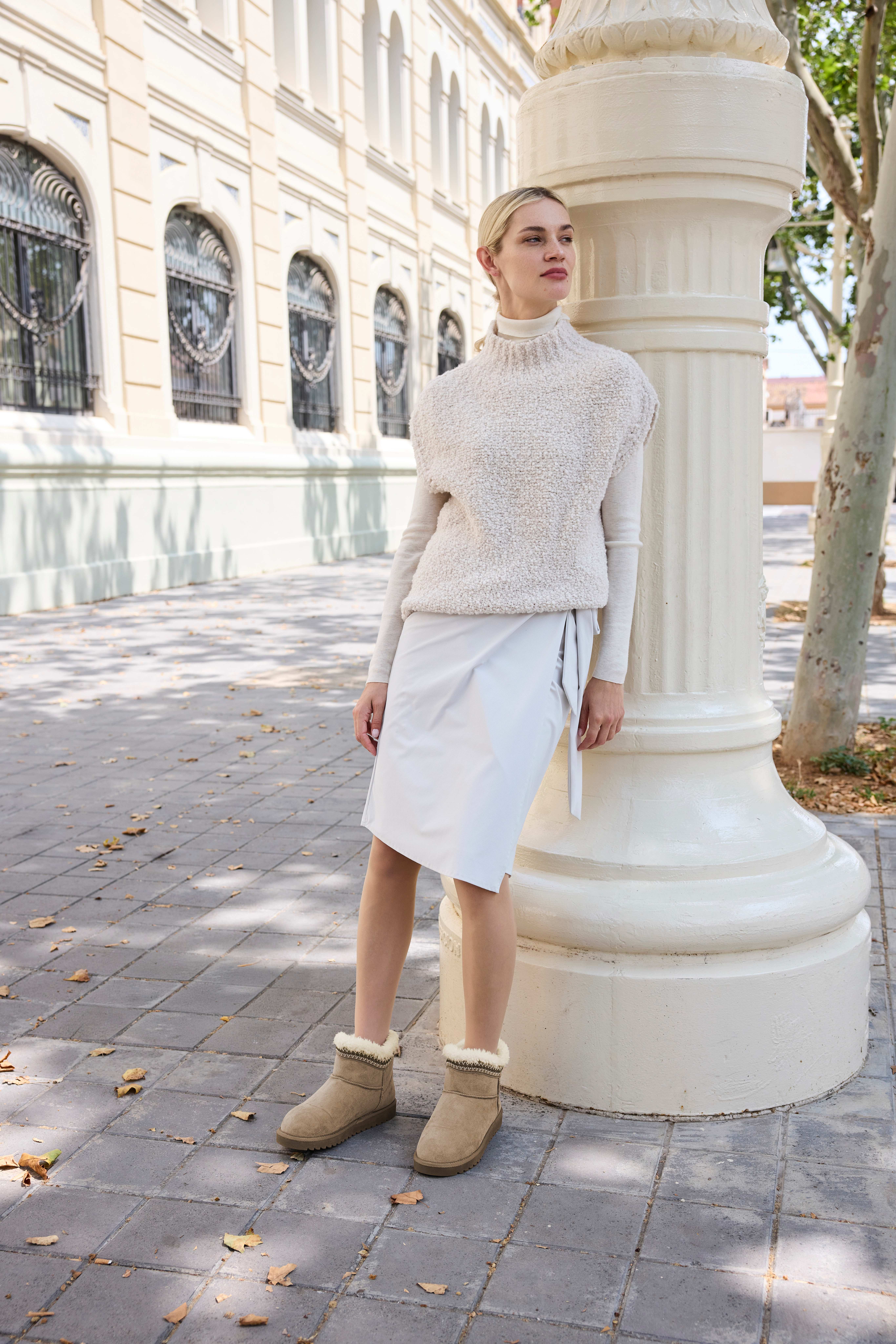 Blonde Frau in weißem Outfit, lehnt an Säule, trägt Australien-Style-Boots